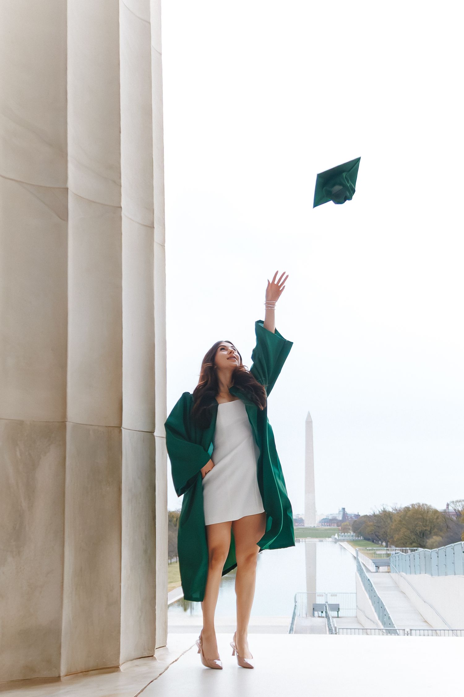 Woman in green graduation gown tosses cap at Lincoln Memorial with Washington Monument in background.