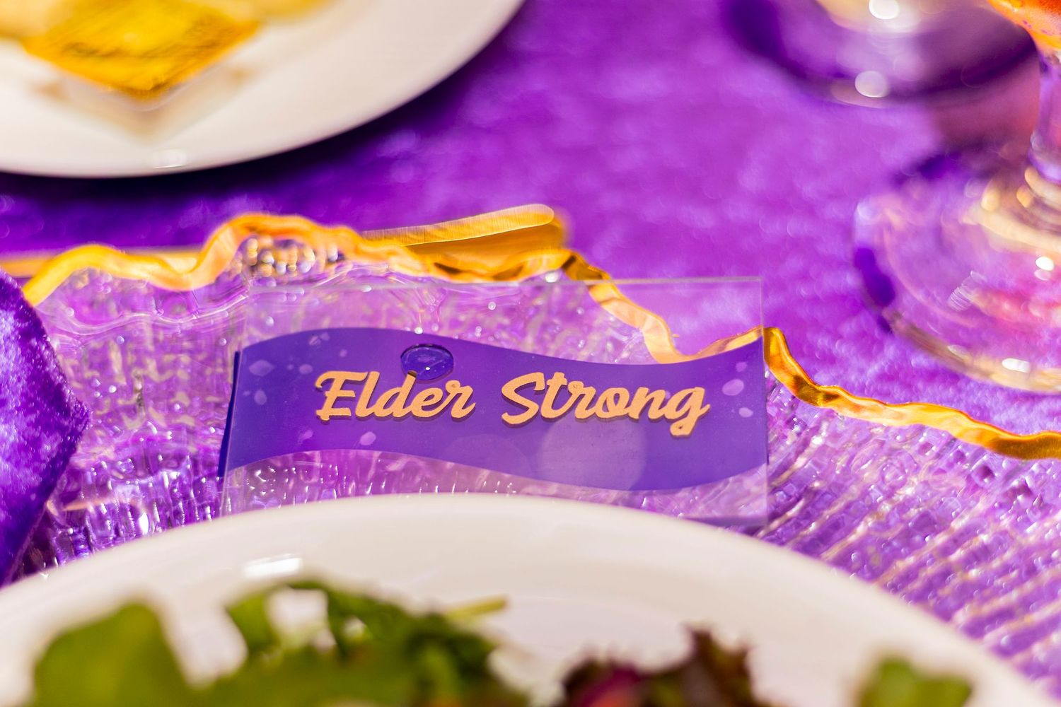 Purple table setting with gold-trimmed place card reading Elder Strong at a formal event.