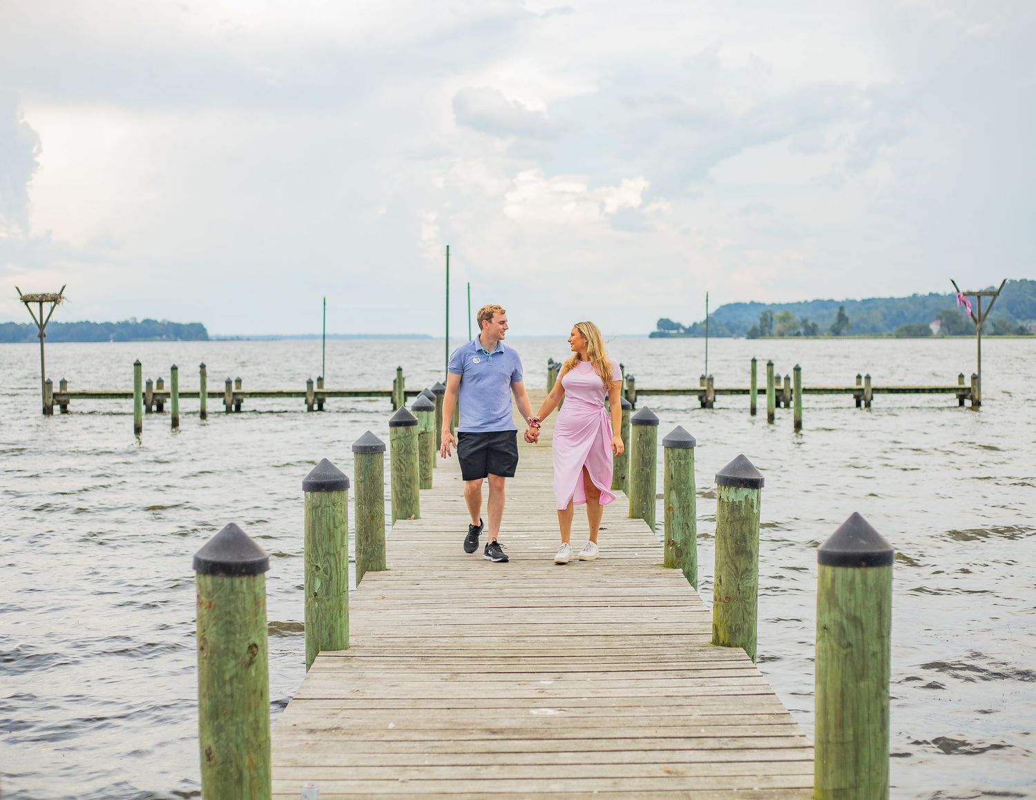 People walking on a wooden pier extending into the water with boats and mountains visible in the distance.