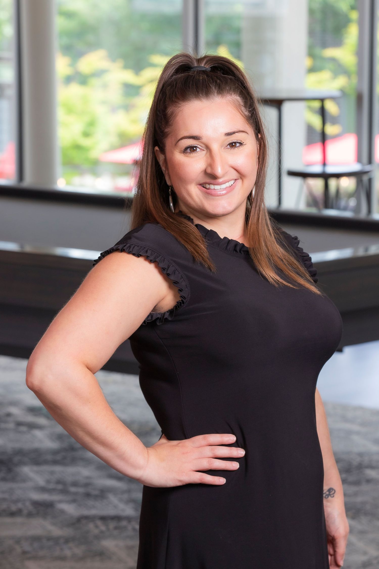 A smiling brunette woman in a black dress poses confidently with hand on hip in a modern well-lit lobby.