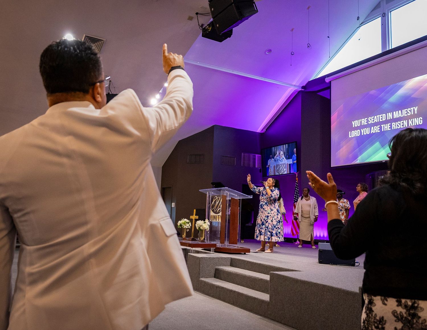 Congregation worshipping in modern church with raised hands, speaker at podium, and praise lyrics displayed on screen.