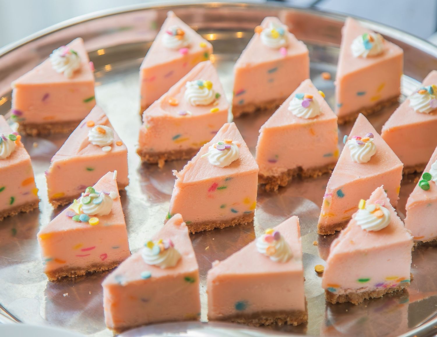 Pink cheesecake slices with sprinkles and whipped cream on a silver tray.
