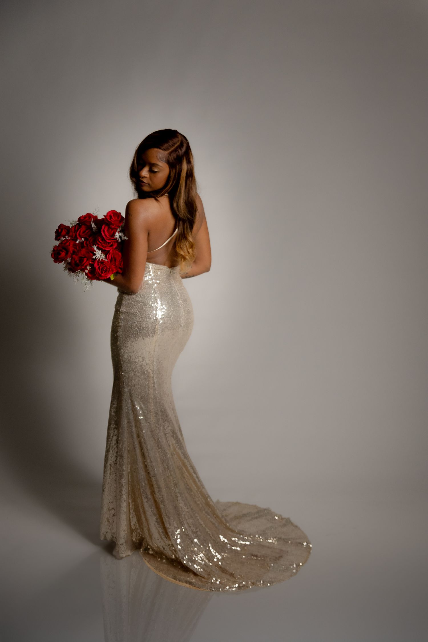 Bride in backless sequin gown holding red rose bouquet, posing elegantly against white background.