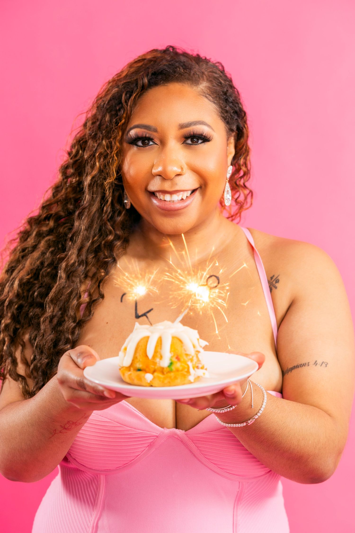 Smiling woman in pink dress holding a bundt cake with sparkler candle against a bright pink background.