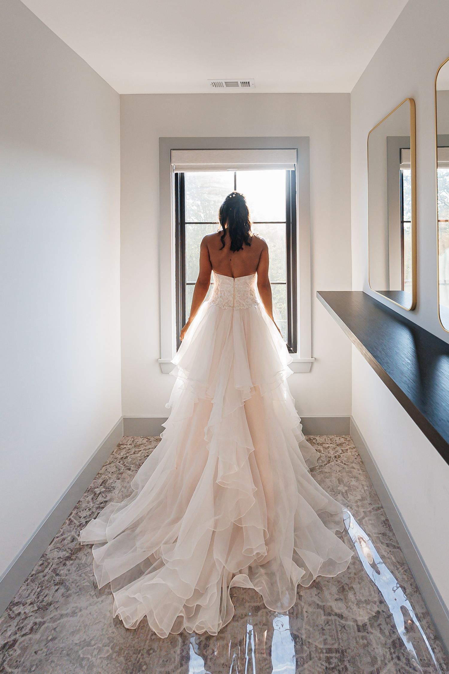 A bride in a flowing white layered wedding gown stands in a hallway looking out a window.
