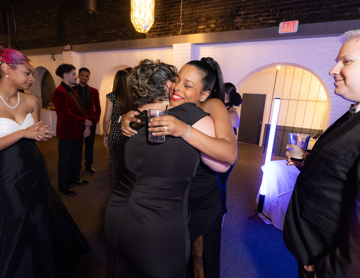 Two women in black dresses share a warm hug at an elegant evening event with guests in formal attire.