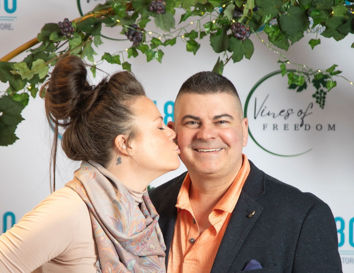 A woman kisses a smiling man on the cheek at a Vines of Freedom event backdrop.