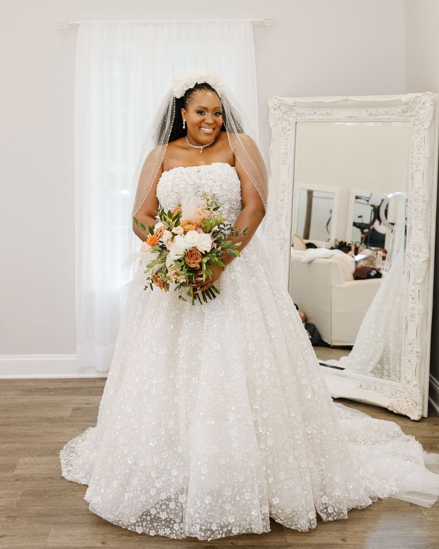 Full-length view of a lace wedding gown with floral bouquet in an elegant white room setting.