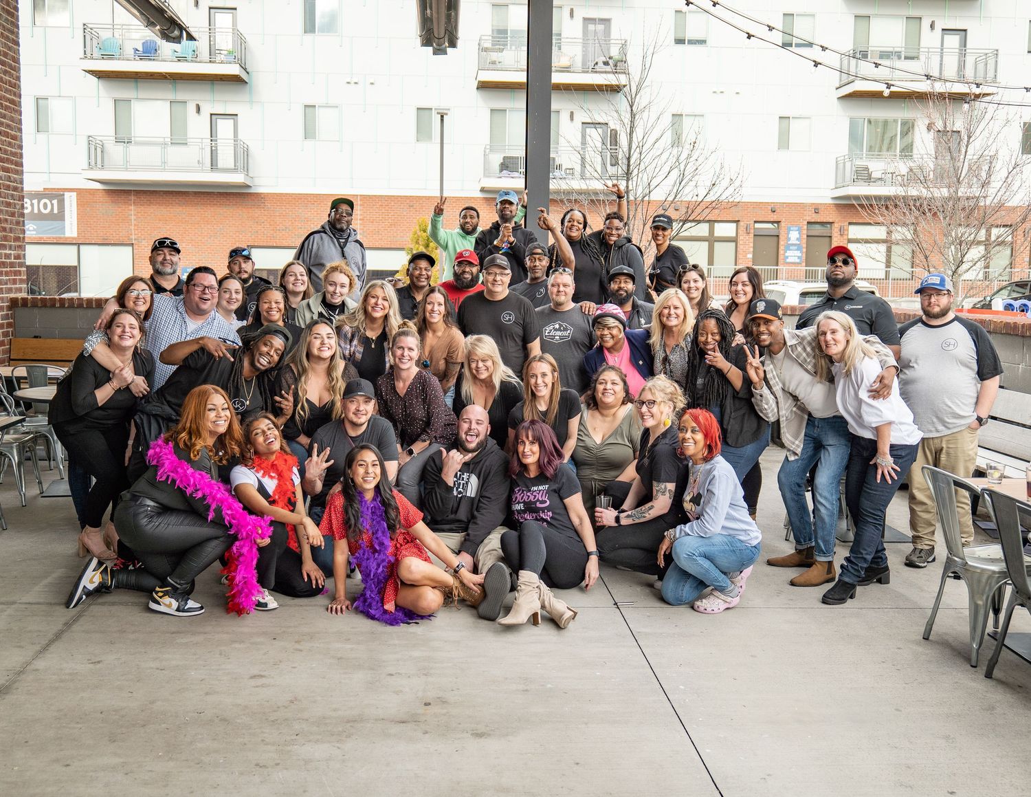 Large group of people posing together outdoors at a patio event, smiling and celebrating in casual attire.