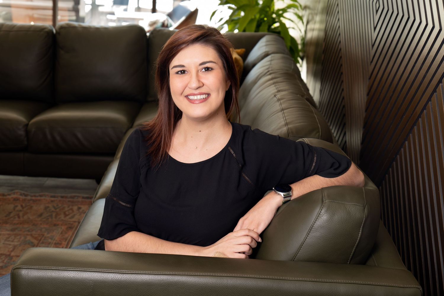 Smiling young woman in black top seated on green leather sofa in modern office lounge area.