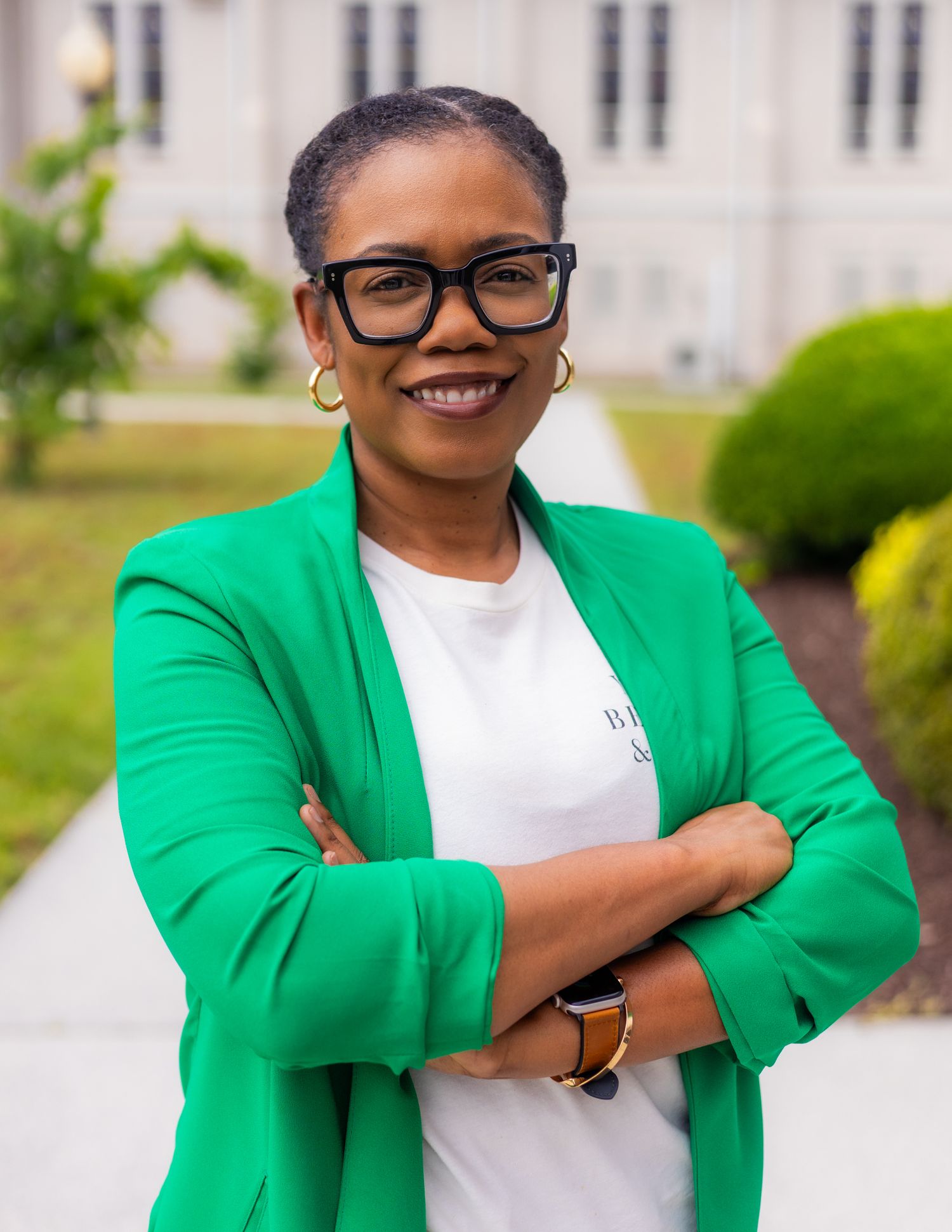 Professional woman in green blazer and glasses smiling with arms crossed outdoors.