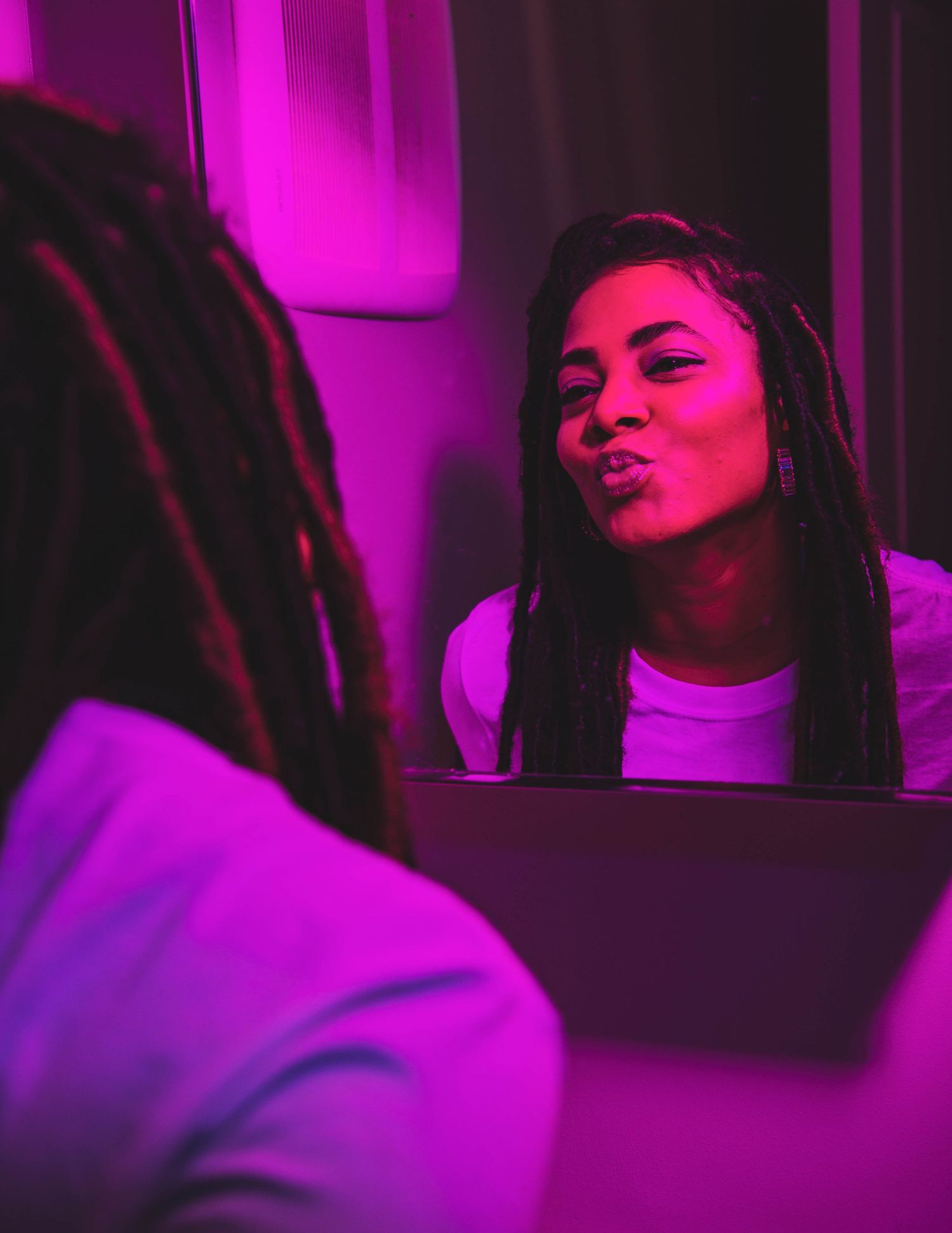 Woman with braids making a kissing face at her reflection in a mirror under pink neon lighting.