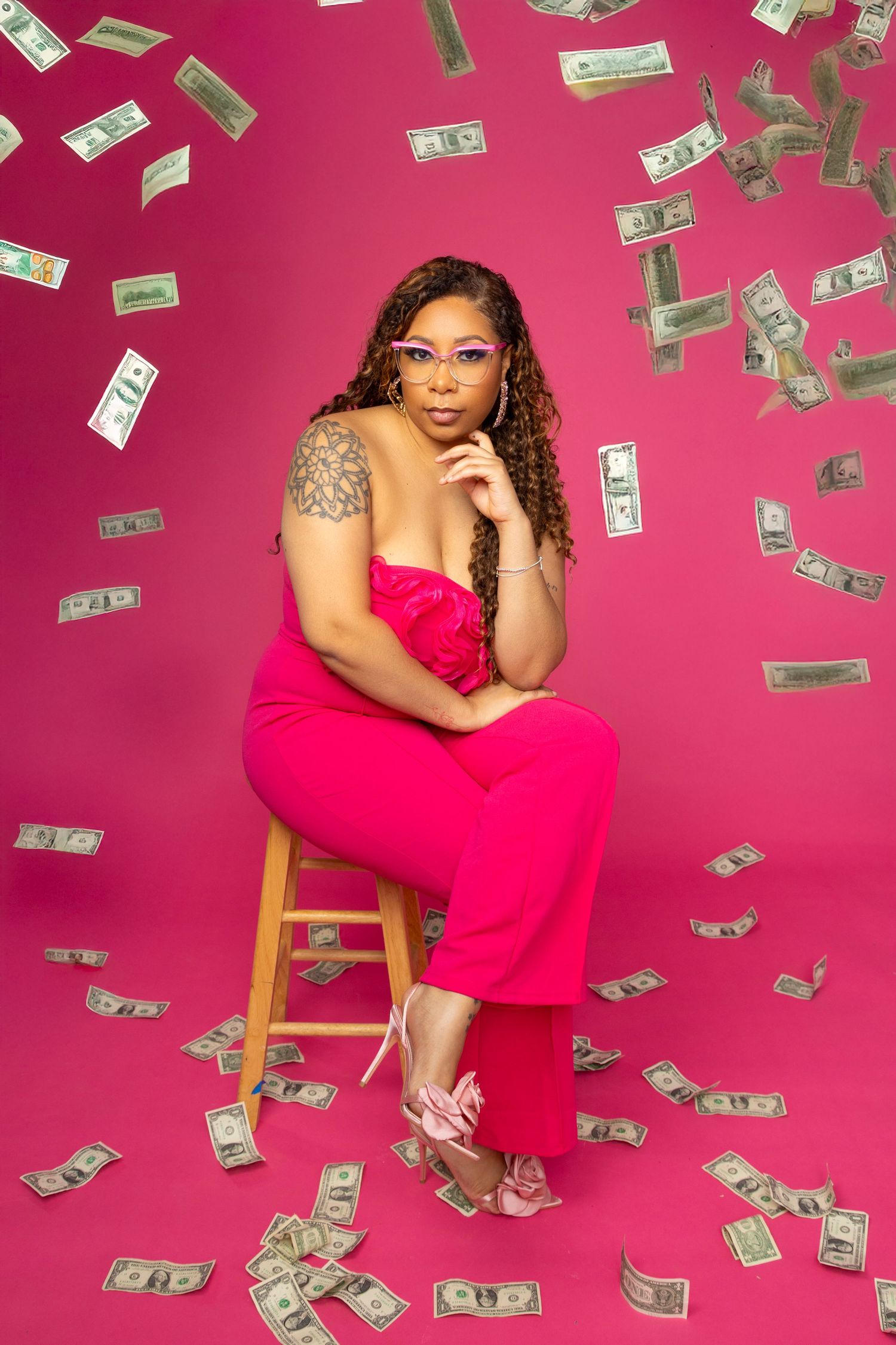 Woman in pink jumpsuit sits on stool surrounded by falling dollar bills against a vibrant pink background.