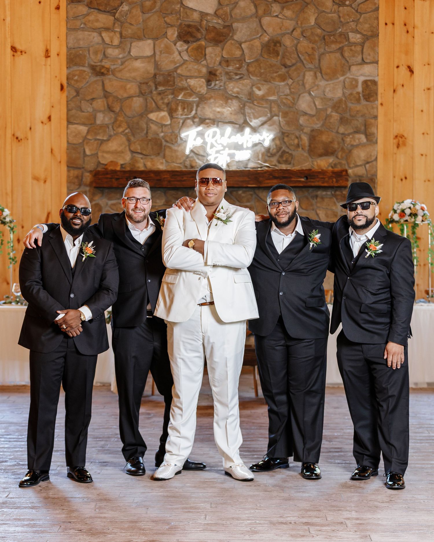 Wedding party in black and white attire posing against a stone wall in formal group photo.