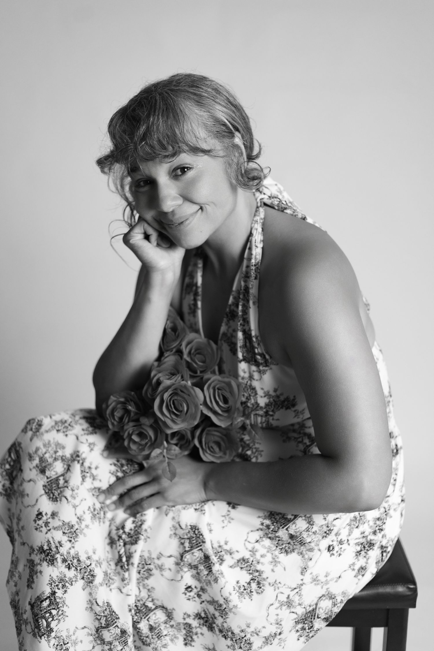 Black and white portrait of a smiling woman in floral dress holding roses, seated with hand on chin.