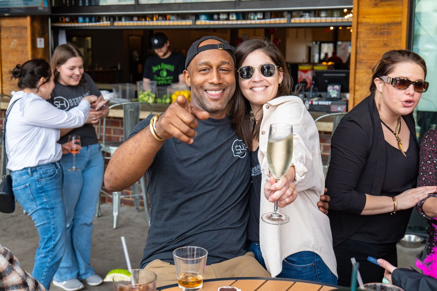 Two friends pose and smile at an outdoor bar patio, one holding a champagne glass, with guests socializing in the background.