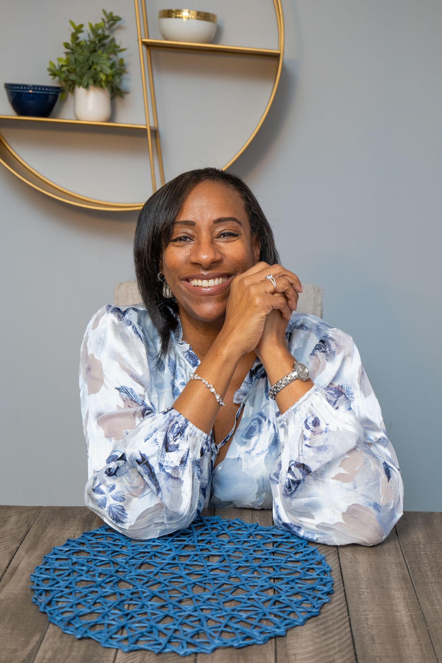 Smiling Black woman in floral blouse seated at wooden table with blue placemat, hands clasped, gold shelf decor behind her.