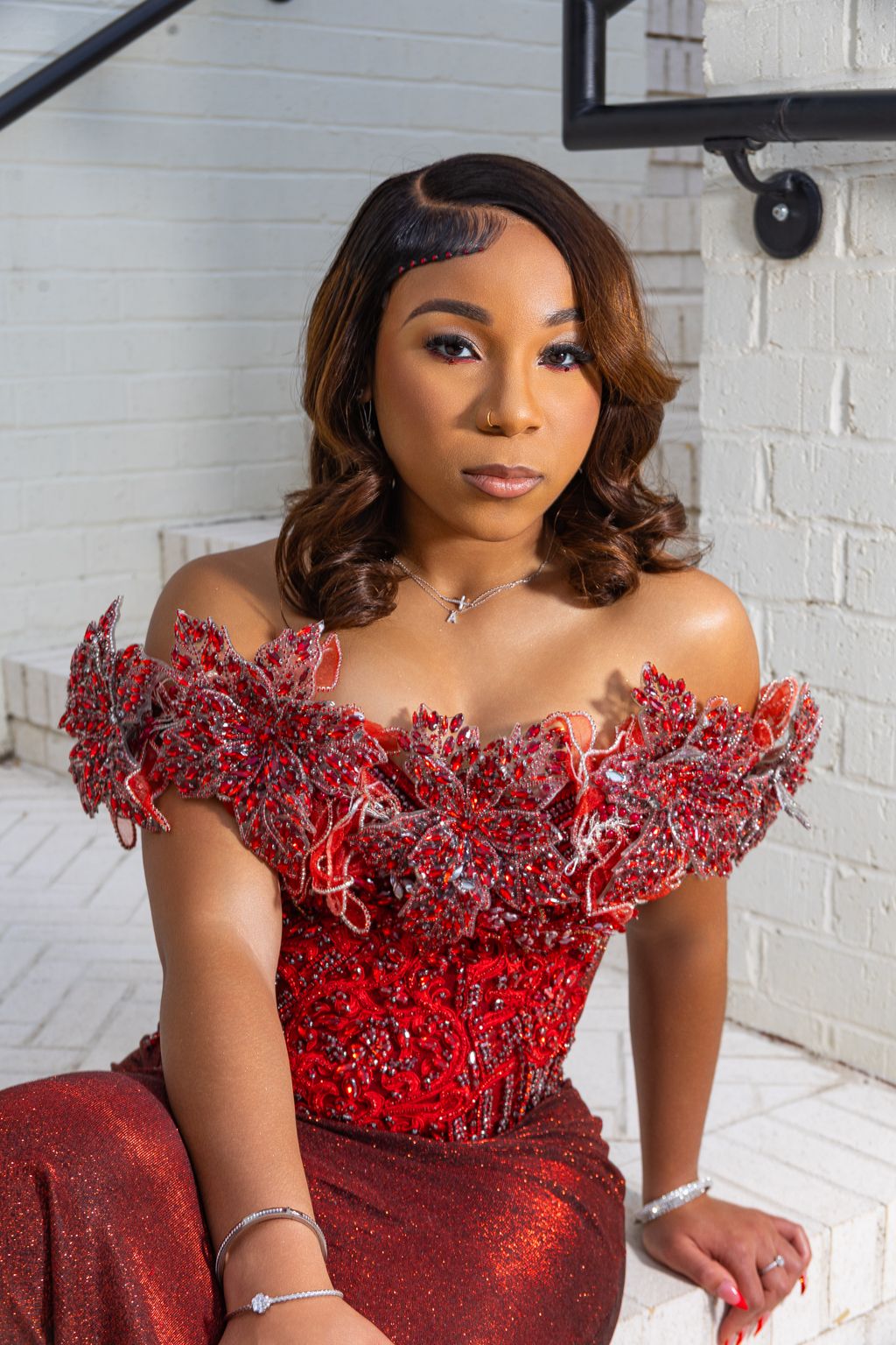 A woman in a red off-shoulder beaded gown with ruffles sits elegantly against a white brick wall.
