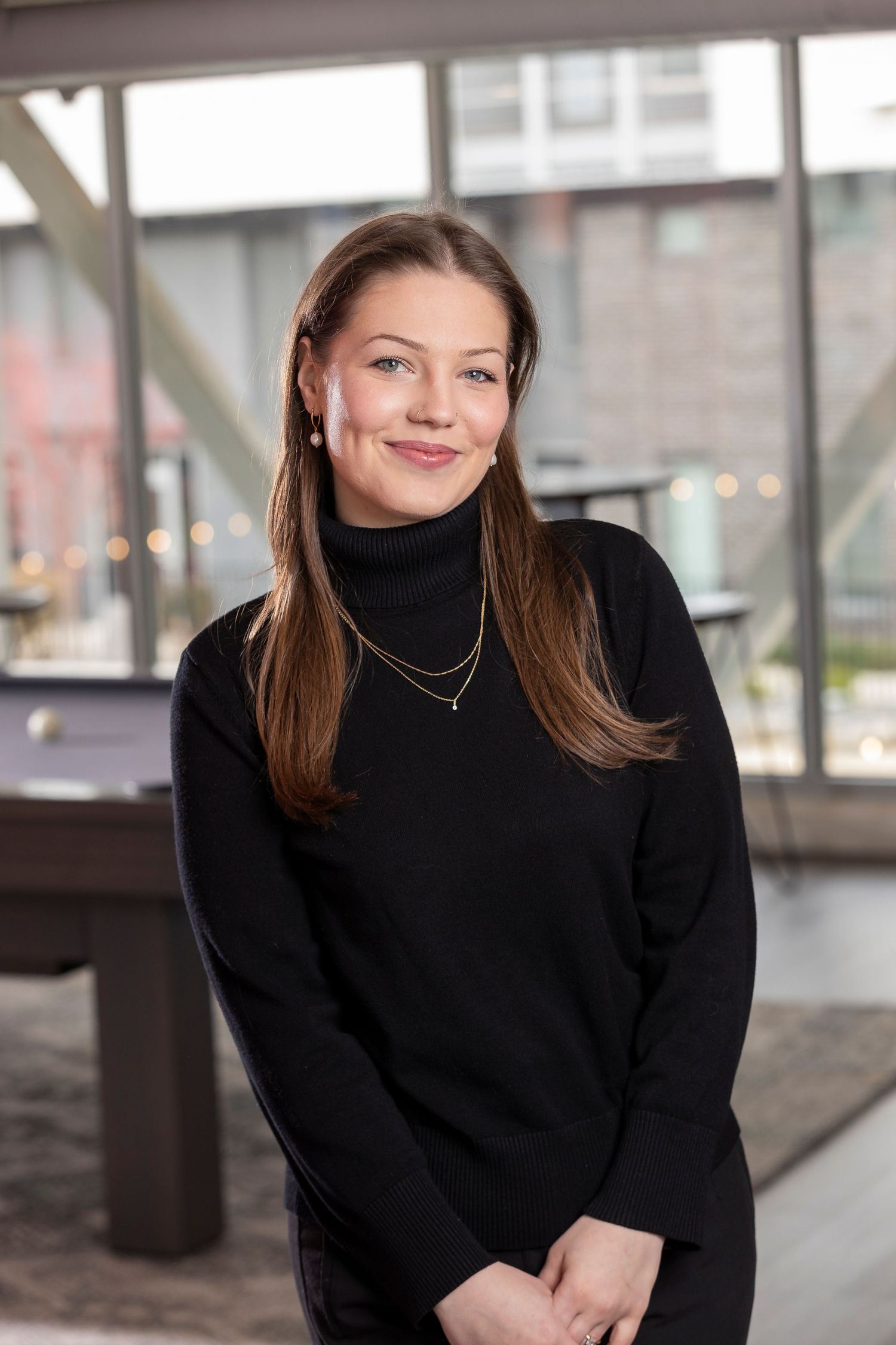 Young woman in black turtleneck sweater with layered necklaces smiling in modern office interior.