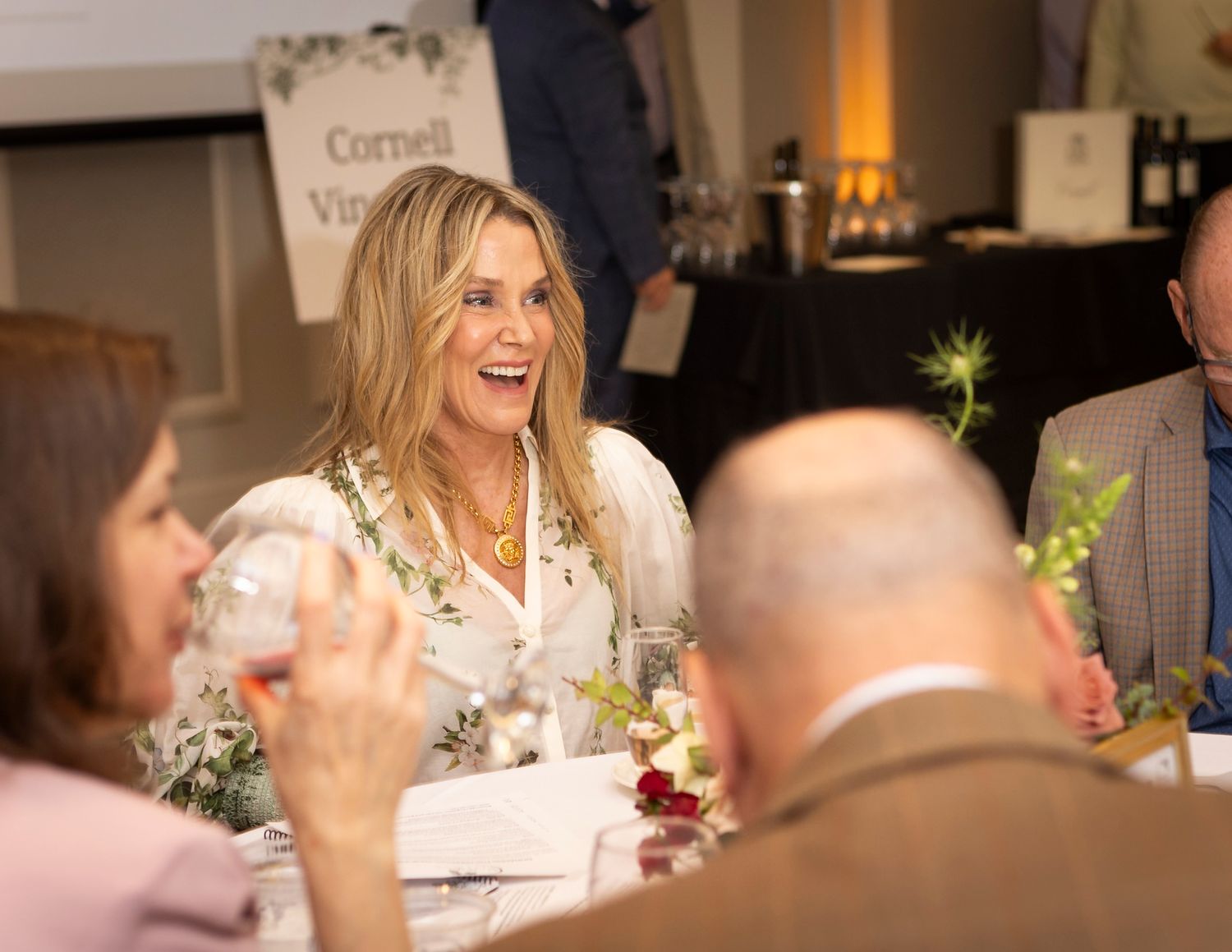 Woman laughing joyfully at an elegant Cornell Vineyard dinner event with guests seated at a floral-decorated table.