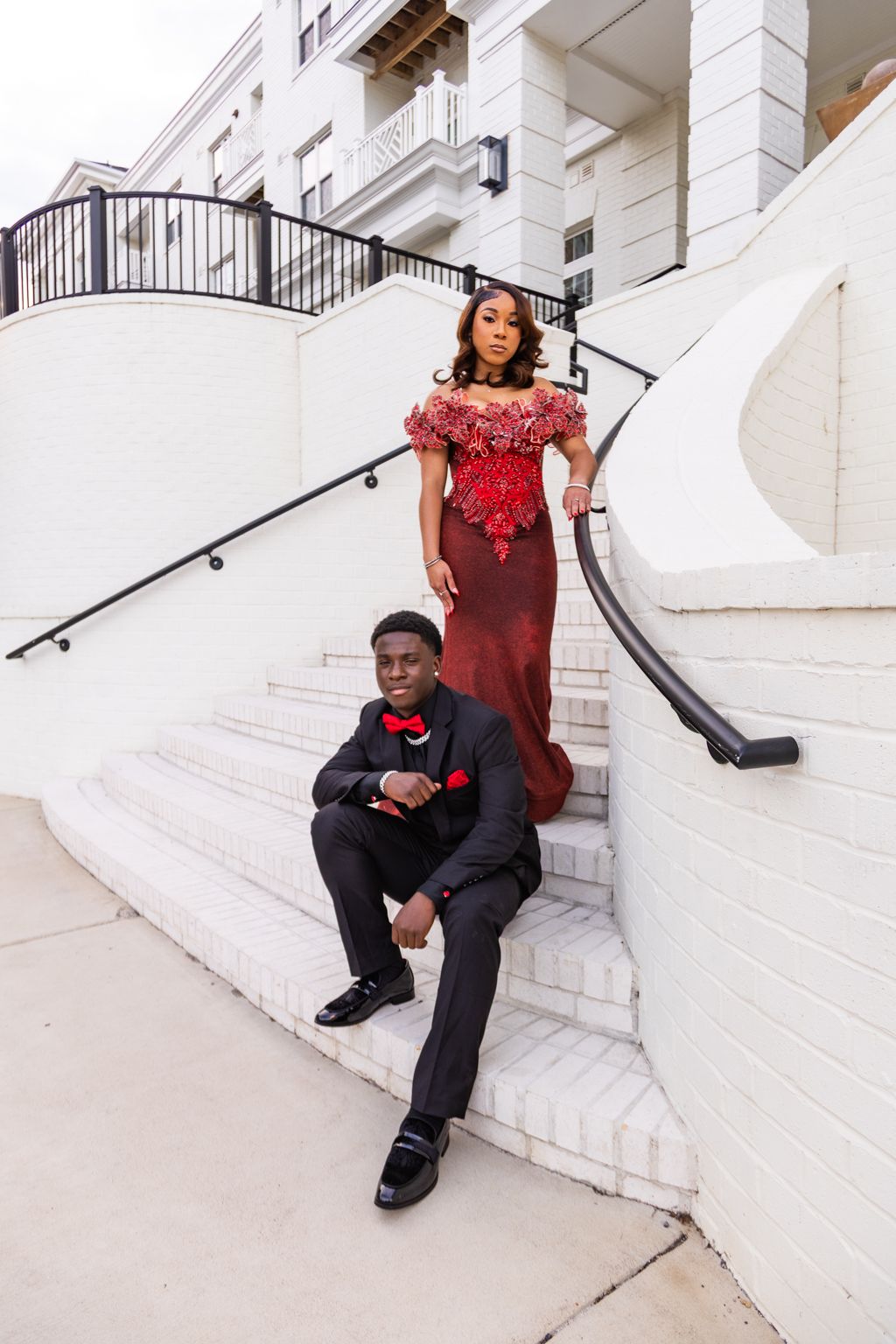 Elegant couple in matching red and black formal wear posing on white outdoor staircase for prom photos.