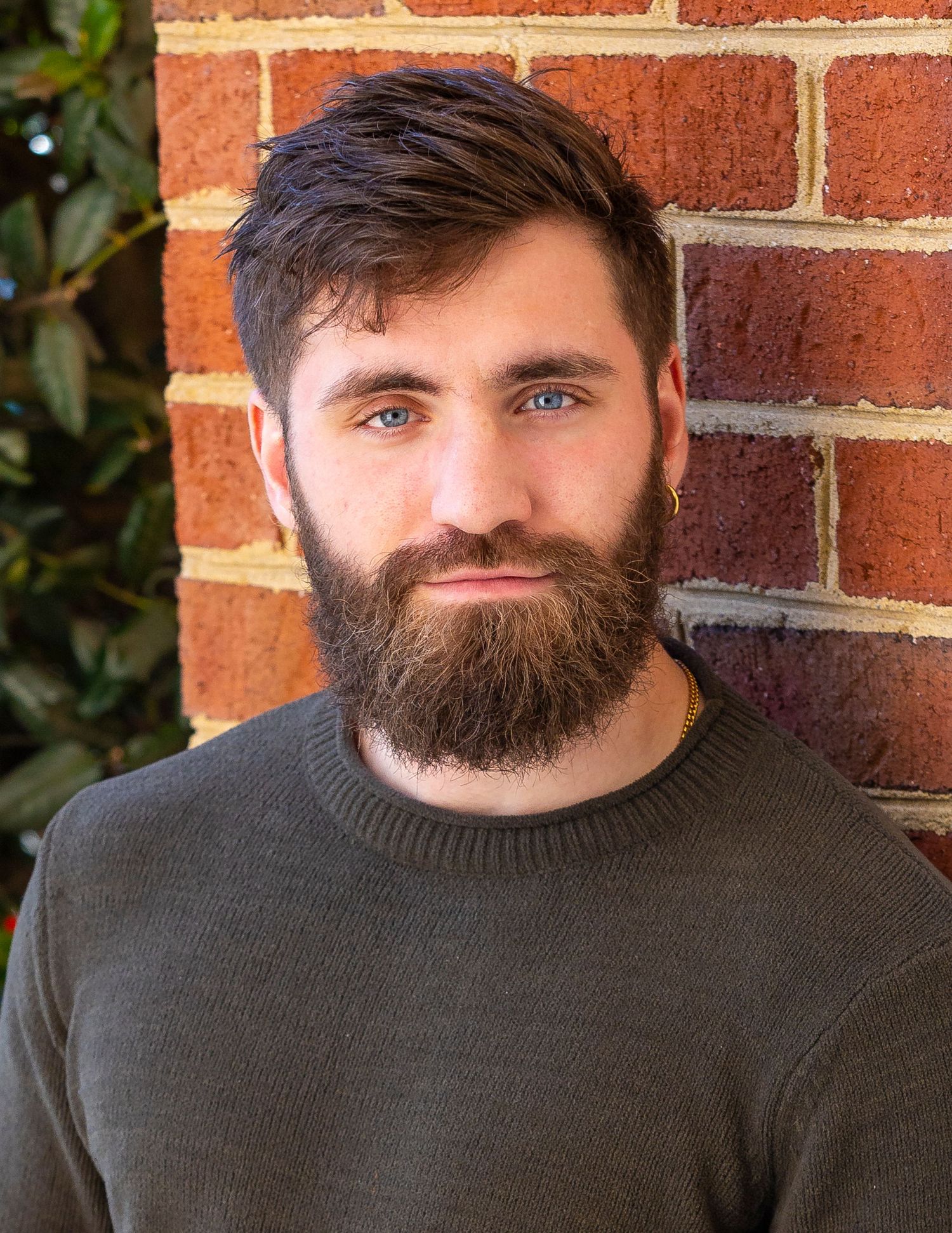 Young bearded man with blue eyes wearing a dark green sweater, posing against a red brick wall outdoors.