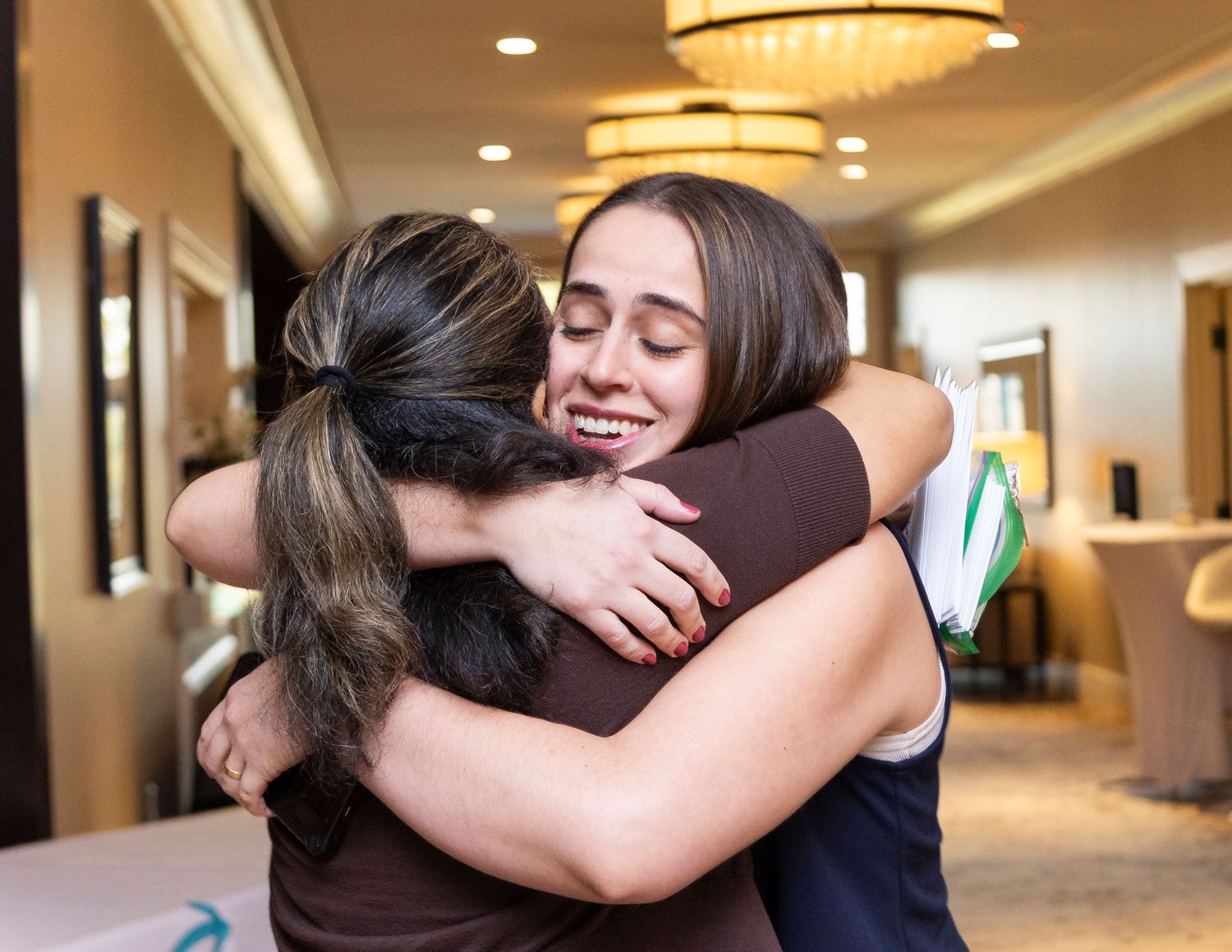 Two women sharing a warm embrace and smiling in an elegant hotel lobby setting.