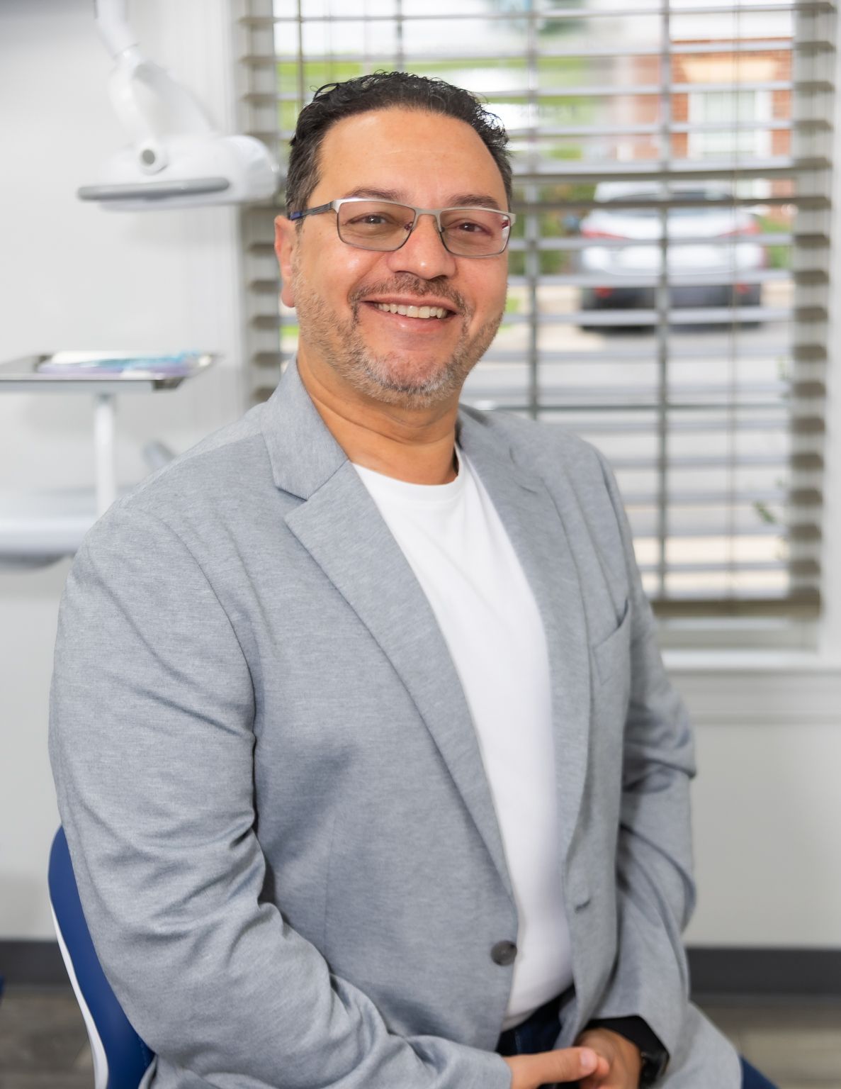 Smiling man in gray blazer and glasses sitting in a modern dental office environment.