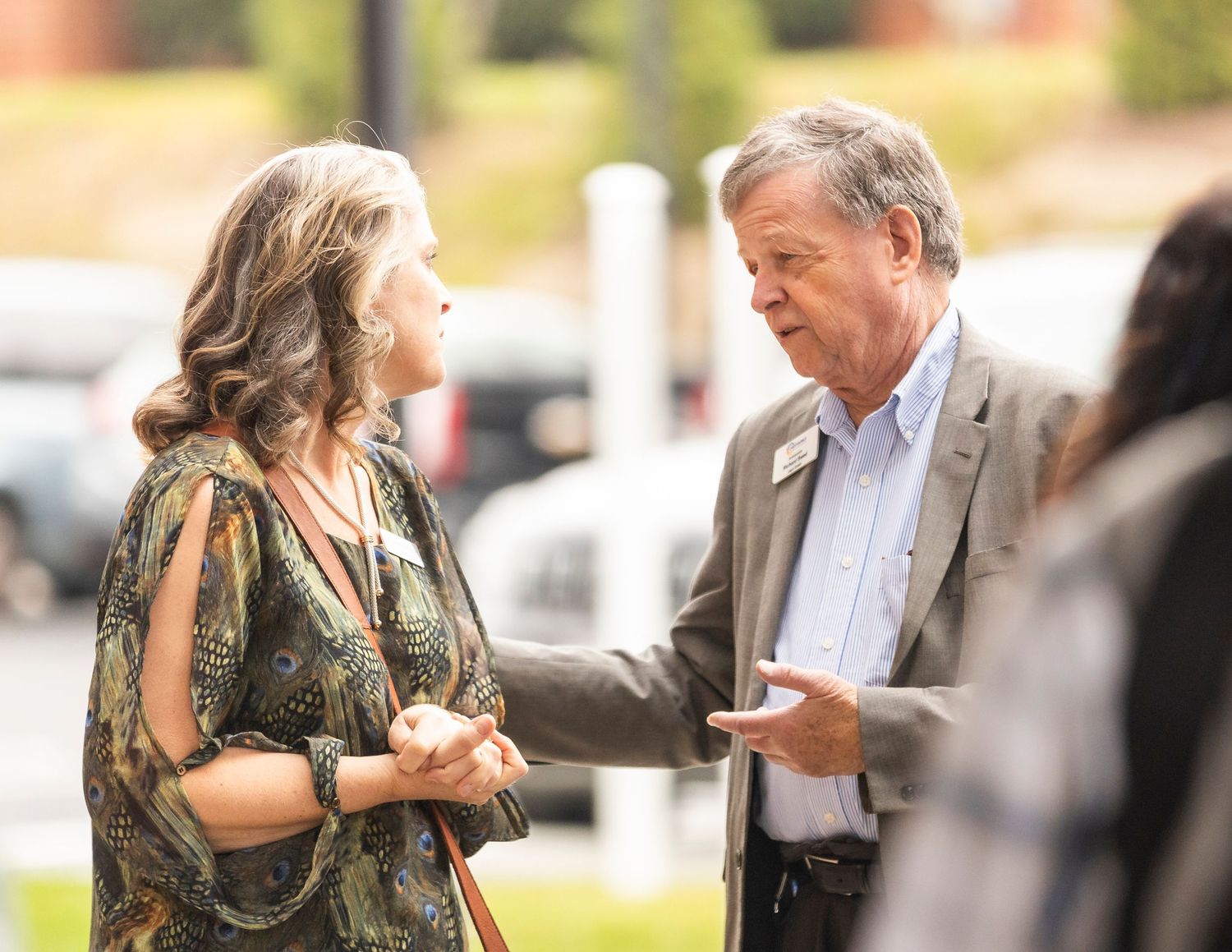 Two people engaged in conversation outdoors, man in blazer gesturing while speaking to woman in floral dress.
