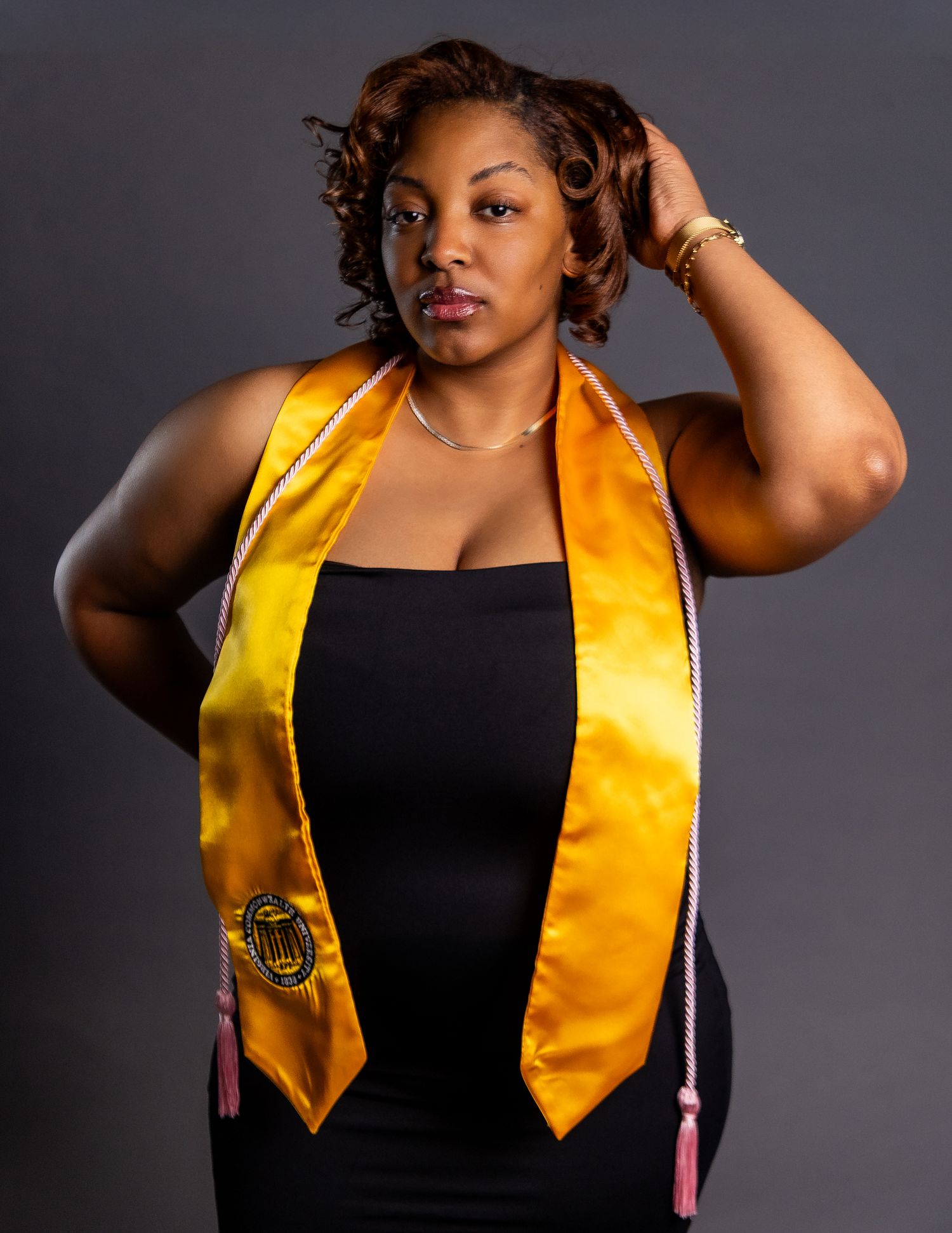 Graduate wearing gold satin stole and pink tassel poses confidently in black dress for senior portrait.
