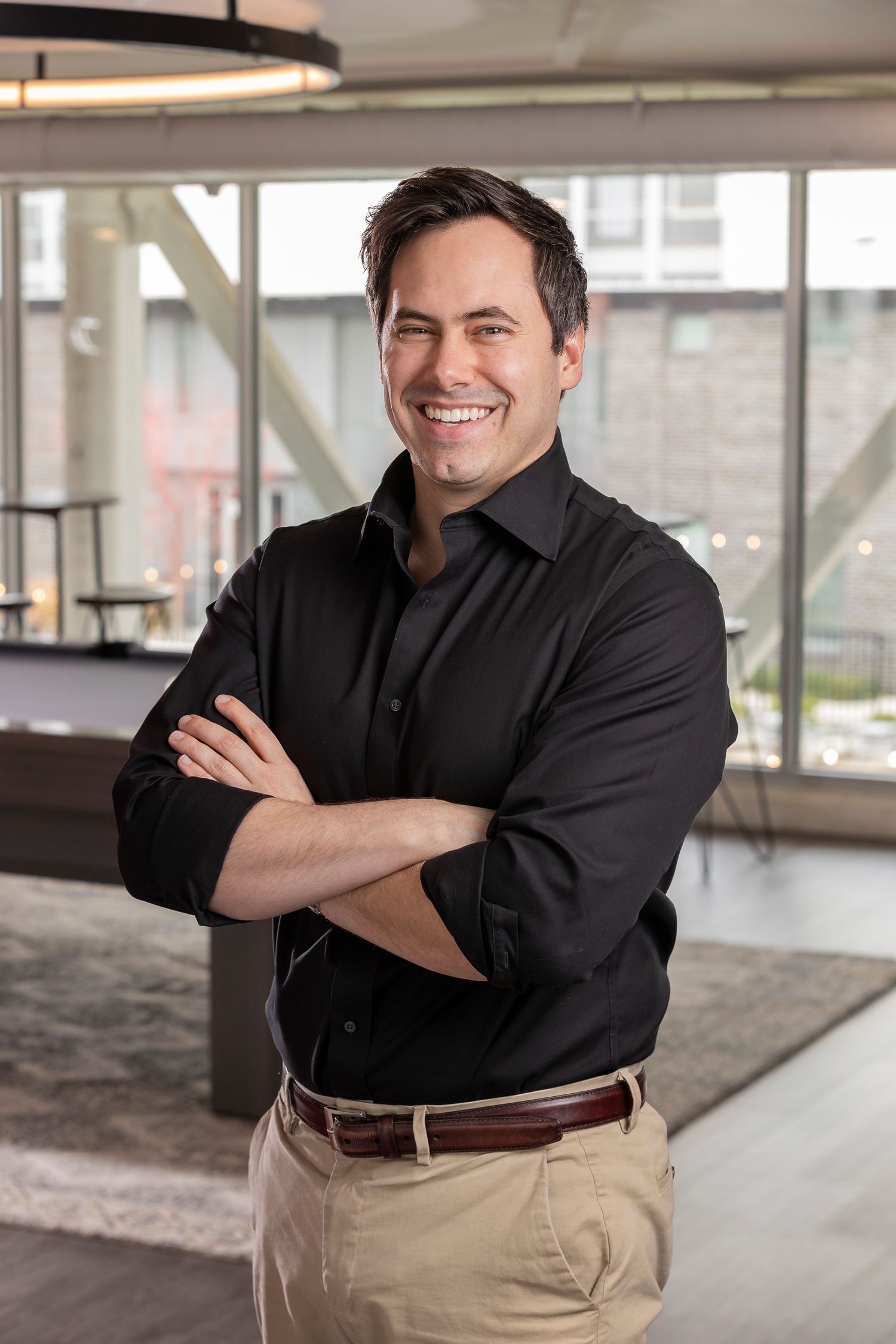 Smiling man in black shirt with arms crossed standing in modern office space with large windows.