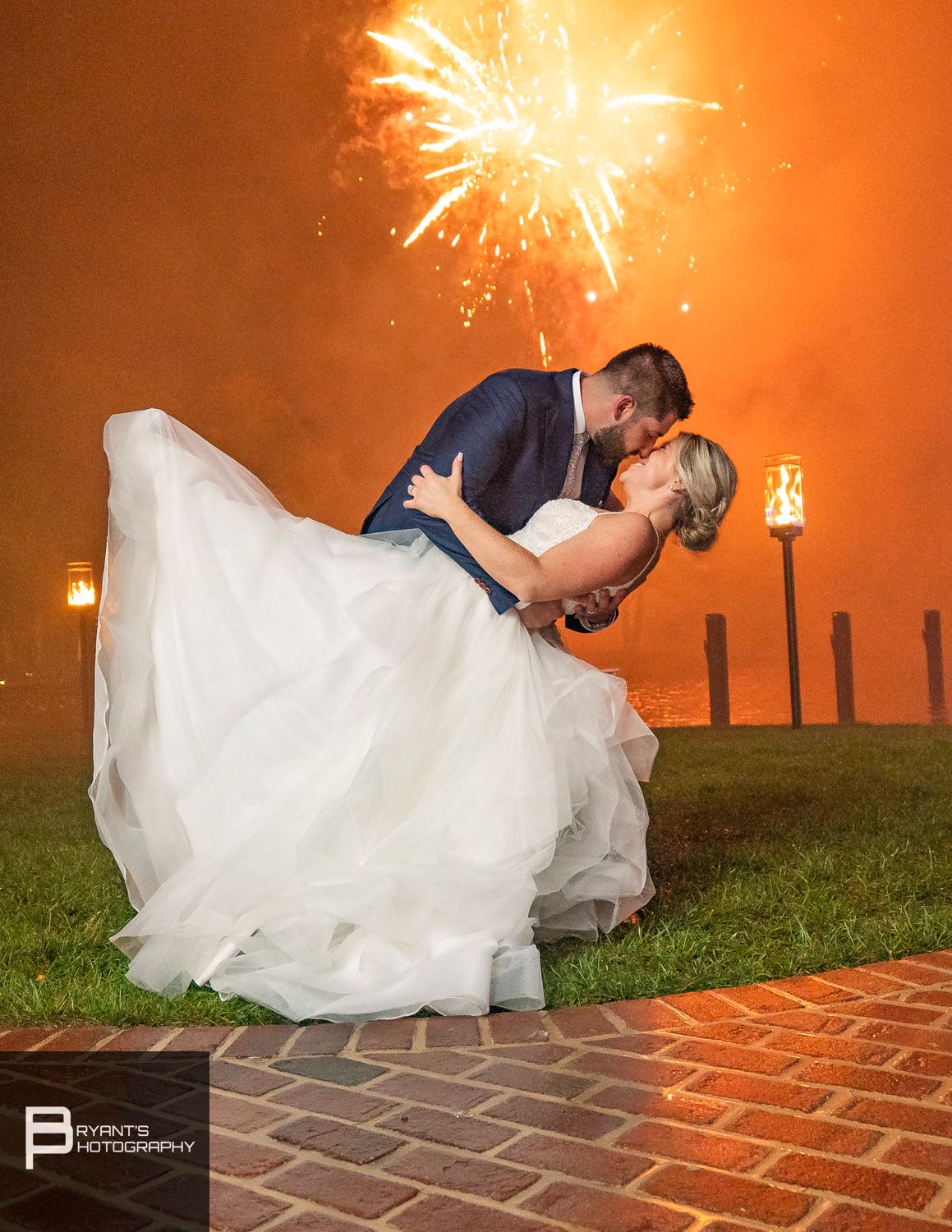 Romantic couple dips and kisses on brick patio with orange fireworks display in background.