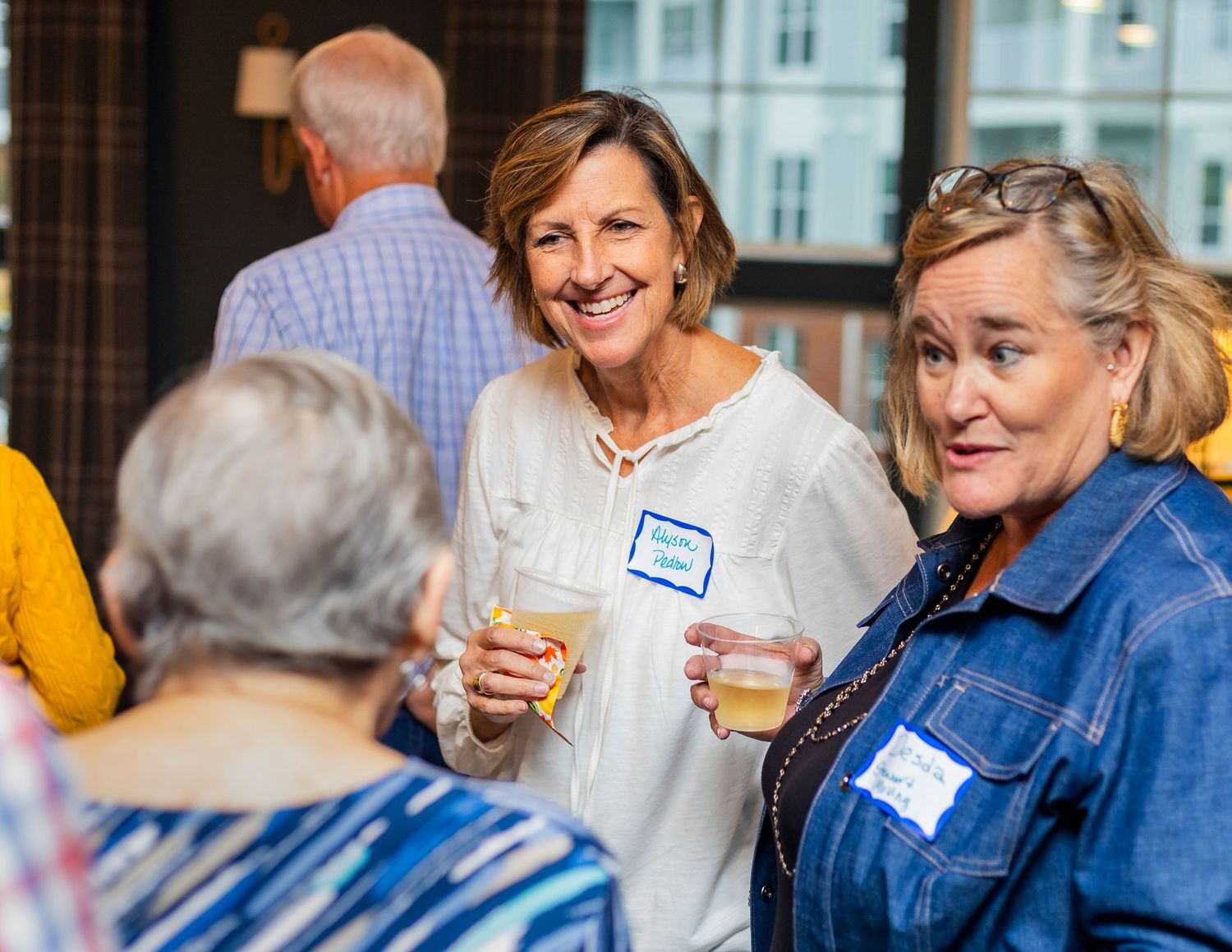 Women socializing at a casual networking event, smiling and holding drinks while wearing name tags indoors.