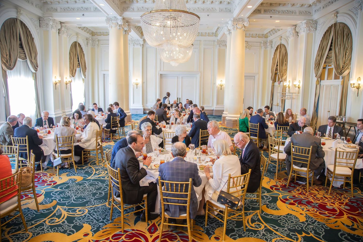Elegant ballroom dining event with guests seated at round tables under chandeliers on ornate carpet.