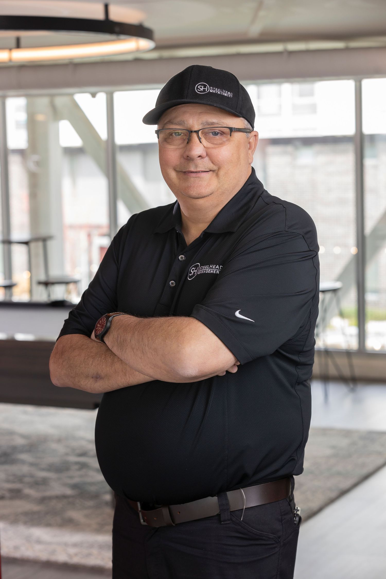 Man in black Nike polo shirt and cap stands with arms crossed, smiling in a modern indoor setting.