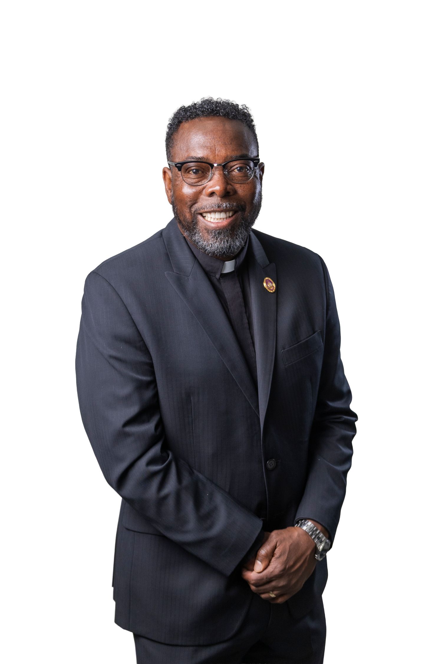 Smiling man in dark suit with clerical collar and glasses posing professionally against white background.