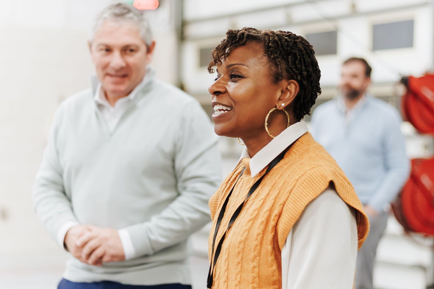 Smiling Black woman in orange vest leading a team discussion in a bright industrial workspace.