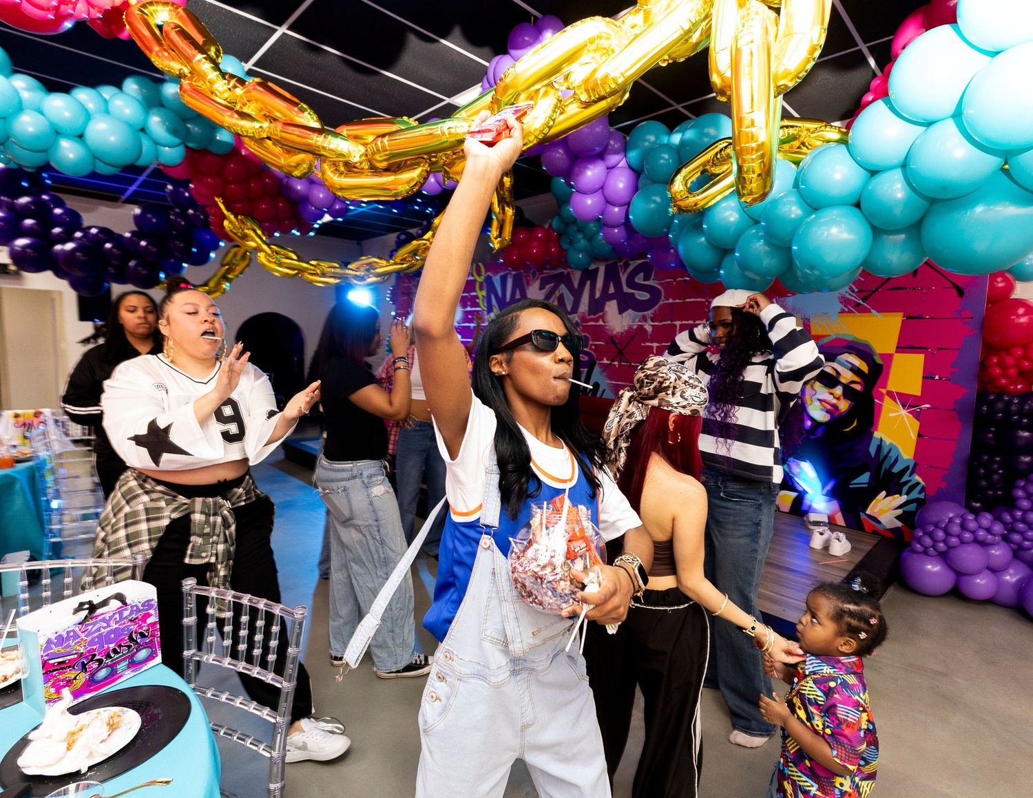 Colorful 90s-themed birthday party with balloon arch decorations, guests dancing and celebrating in vibrant retro outfits.