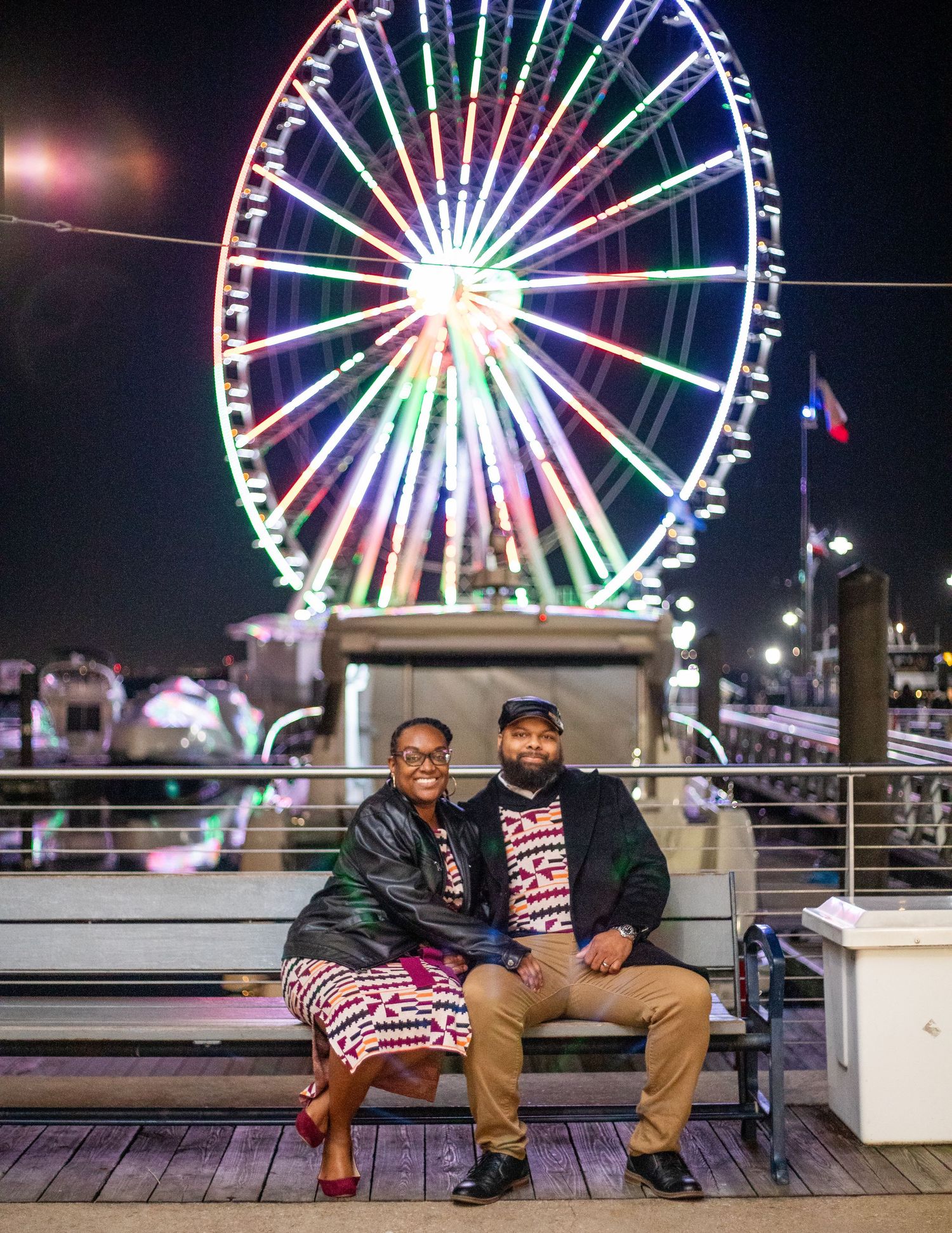 Two people sit on a bench with a brilliantly lit Ferris wheel glowing behind them.