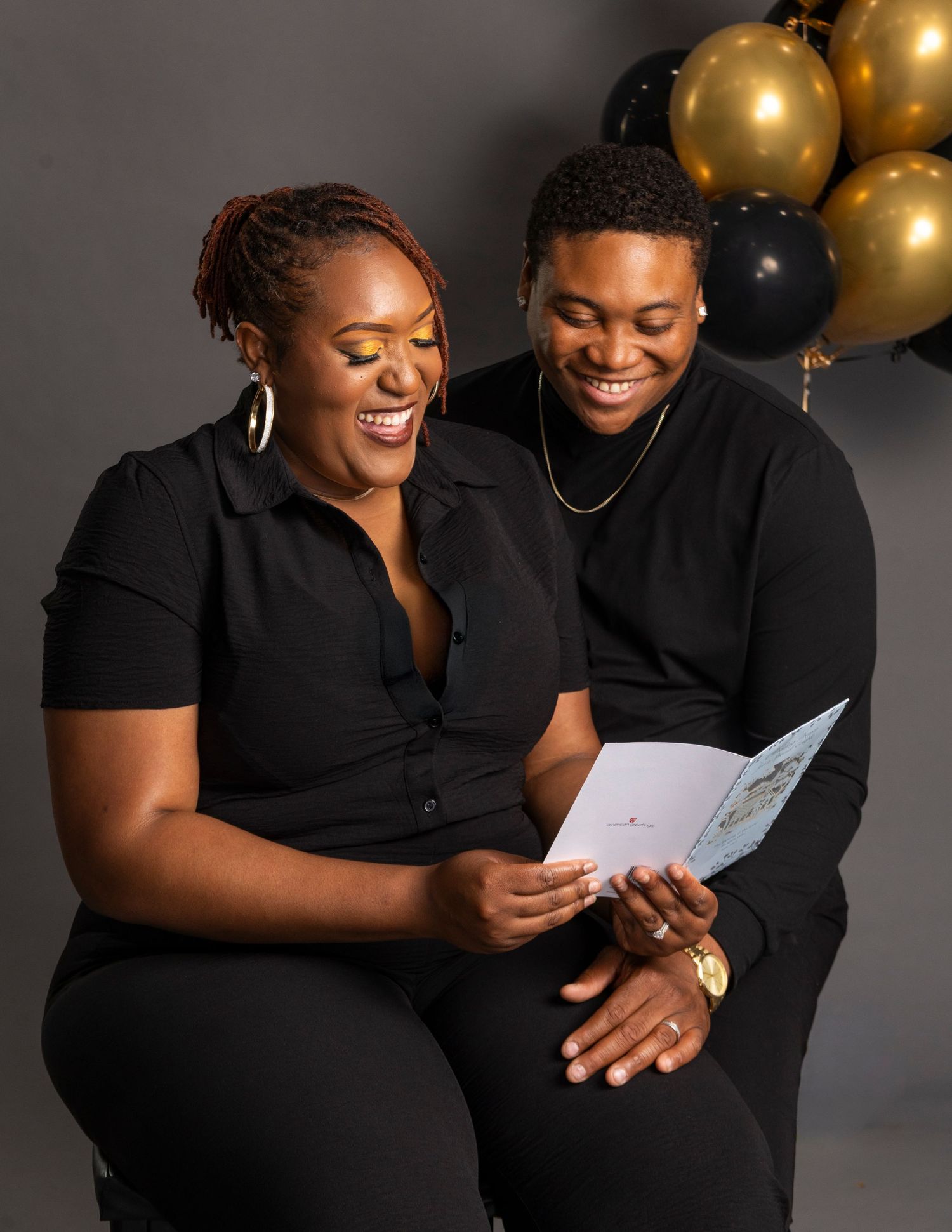 Two people in black outfits smile while reading a greeting card, with gold and black balloons in the background.