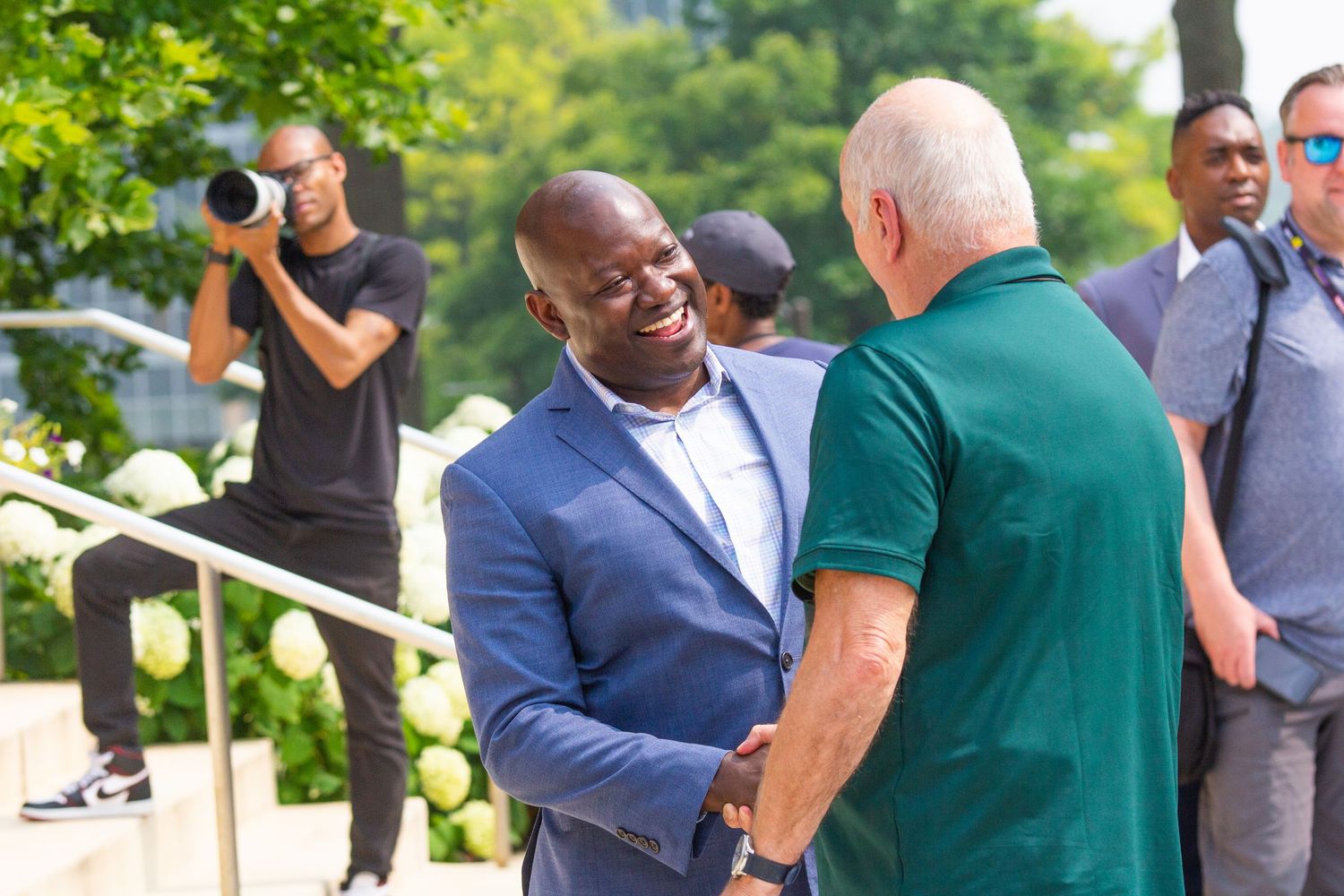 Two men shaking hands outdoors while a photographer captures the moment in a sunny urban setting.