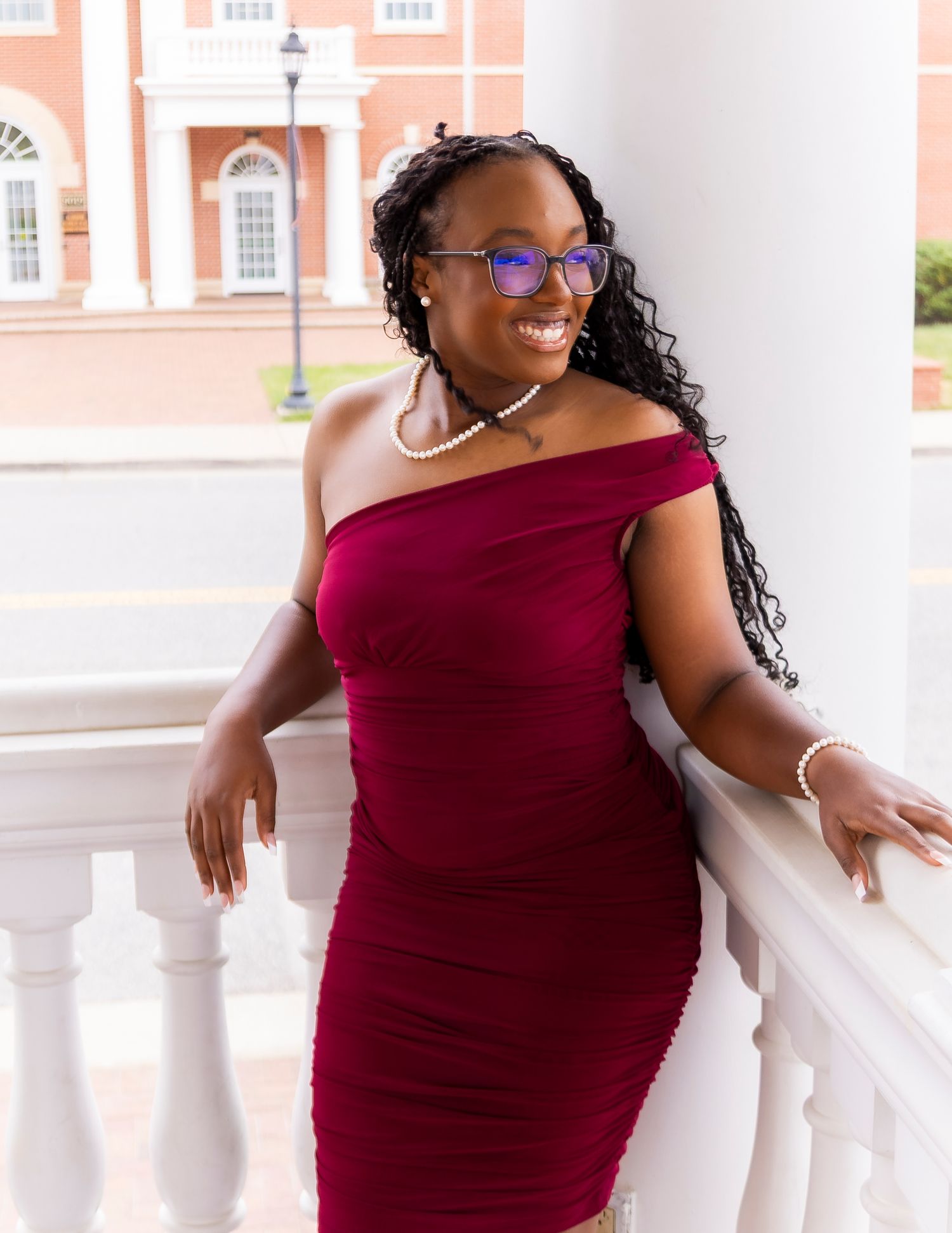 Young woman in burgundy off-shoulder dress poses gracefully on white balcony railing outside elegant brick building.