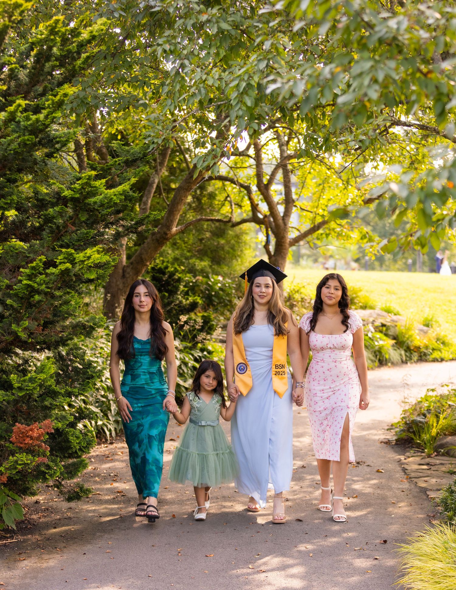 A graduate in cap and gown walks with three family members along a lush garden path on a sunny graduation day.