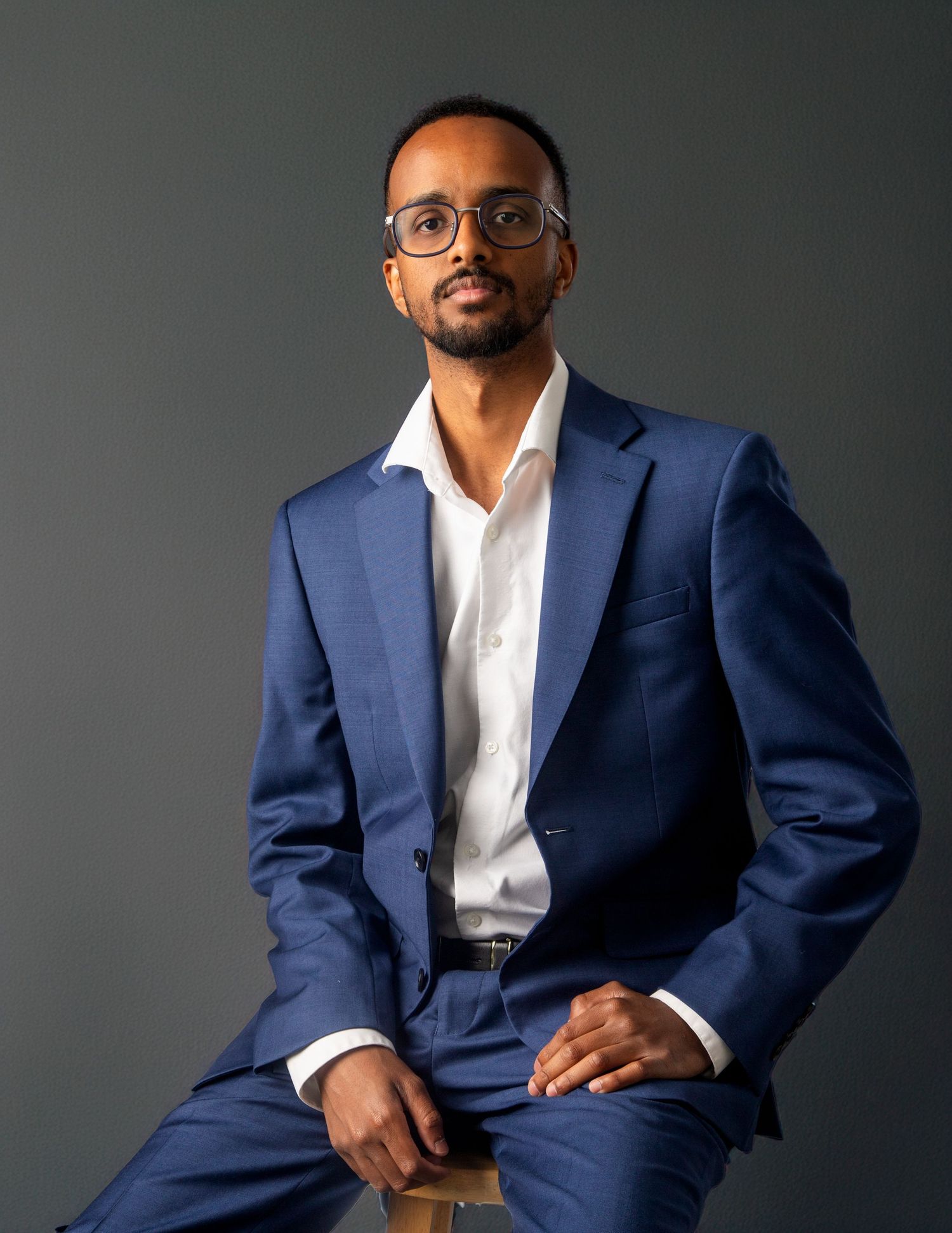 Professional man in blue suit and glasses sitting confidently against gray background for business portrait.