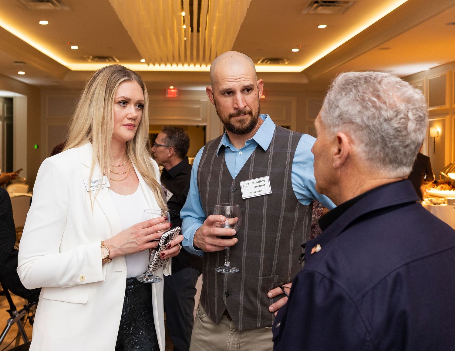 Three professionals networking at a formal event, holding drinks and engaged in conversation in an elegant venue.