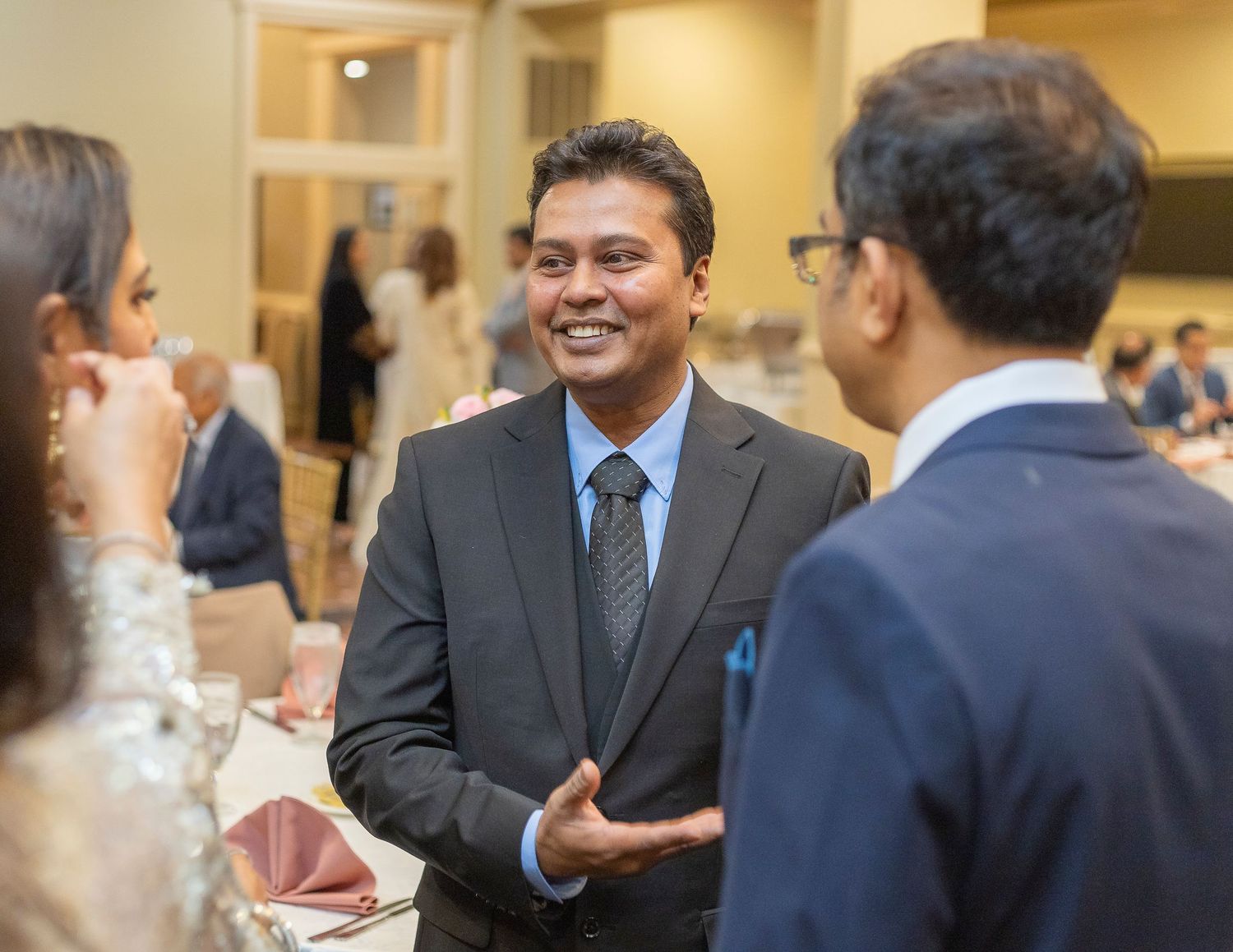 Man in gray suit smiling and conversing at a formal networking event or reception with other guests.