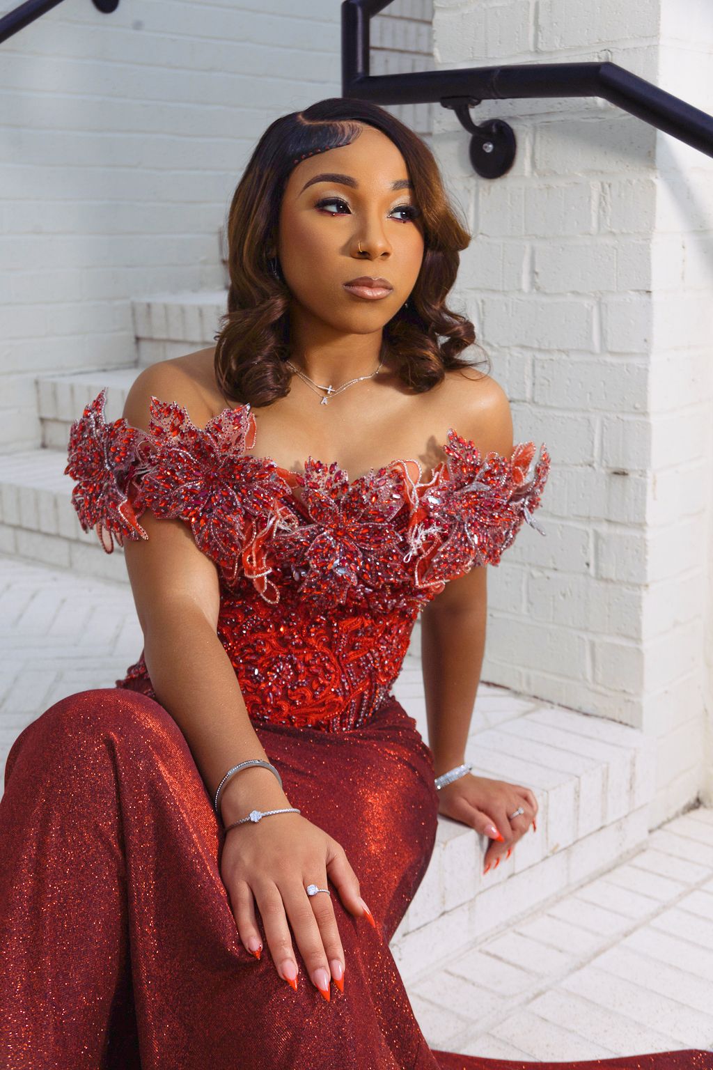 Young woman in a stunning red sequined off-shoulder gown with floral details, posed elegantly on white steps.