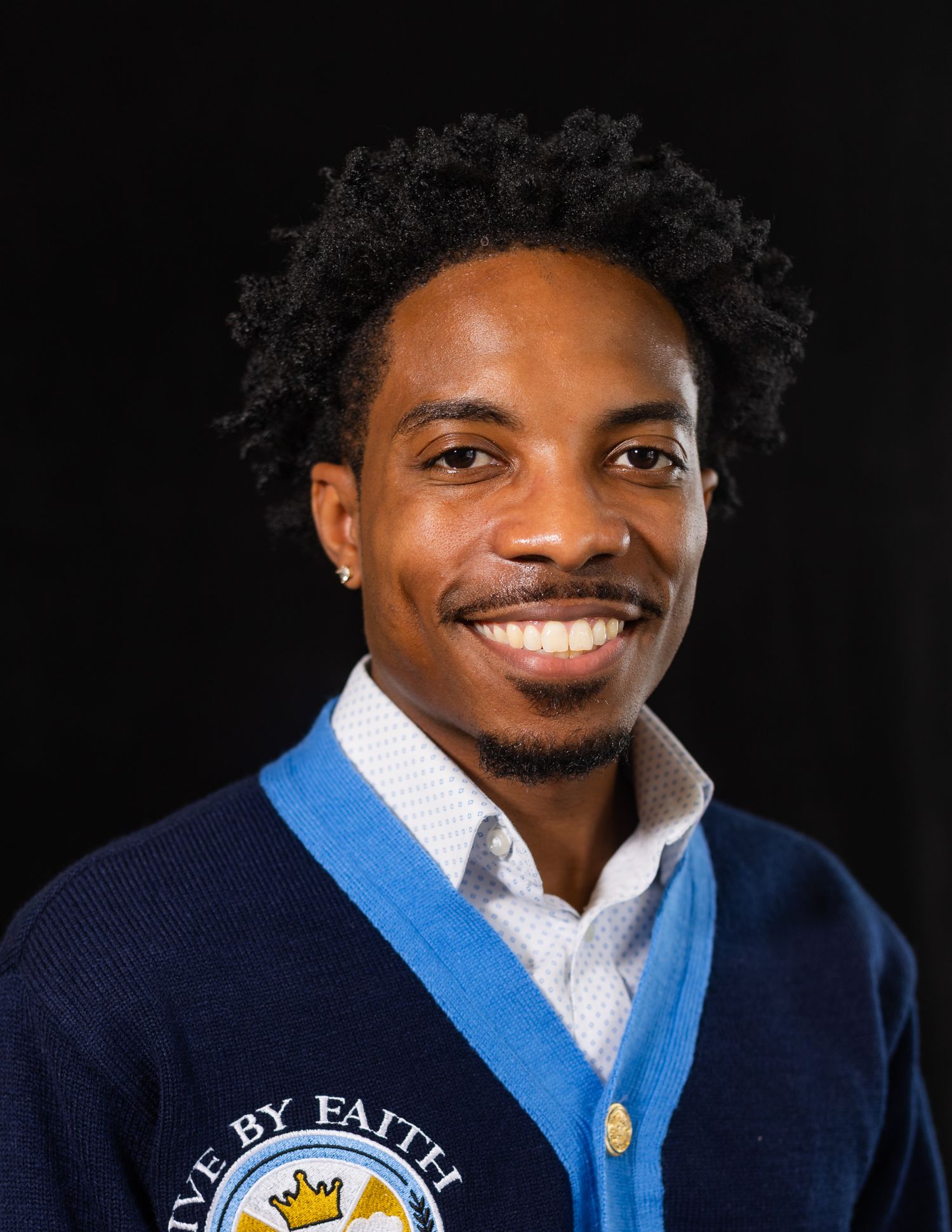 Smiling young man wearing a navy blue cardigan with a Live by Faith logo on a dark background.