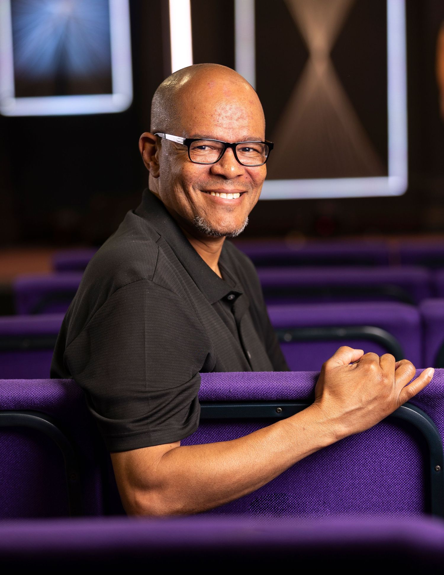 Smiling bald man with glasses wearing black shirt, seated in purple auditorium chairs, posing confidently.