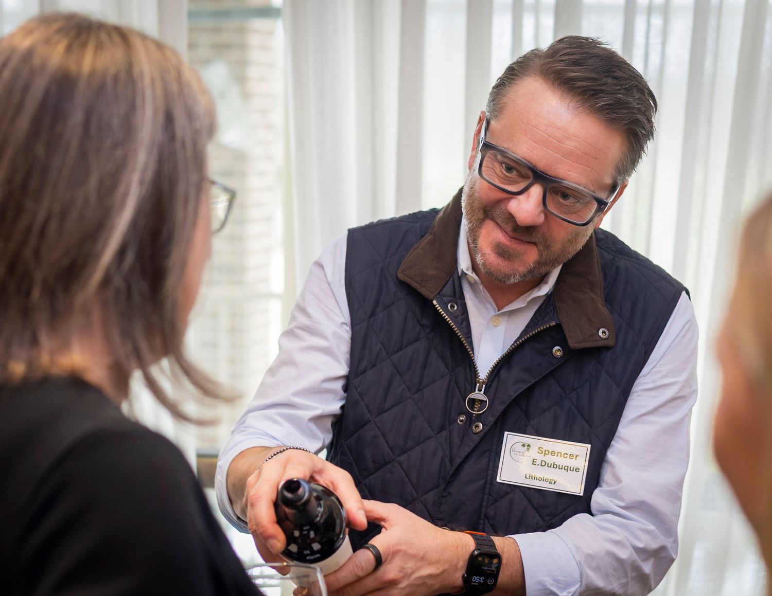 A man in a quilted vest and glasses presents a wine bottle to a woman at an indoor tasting event.